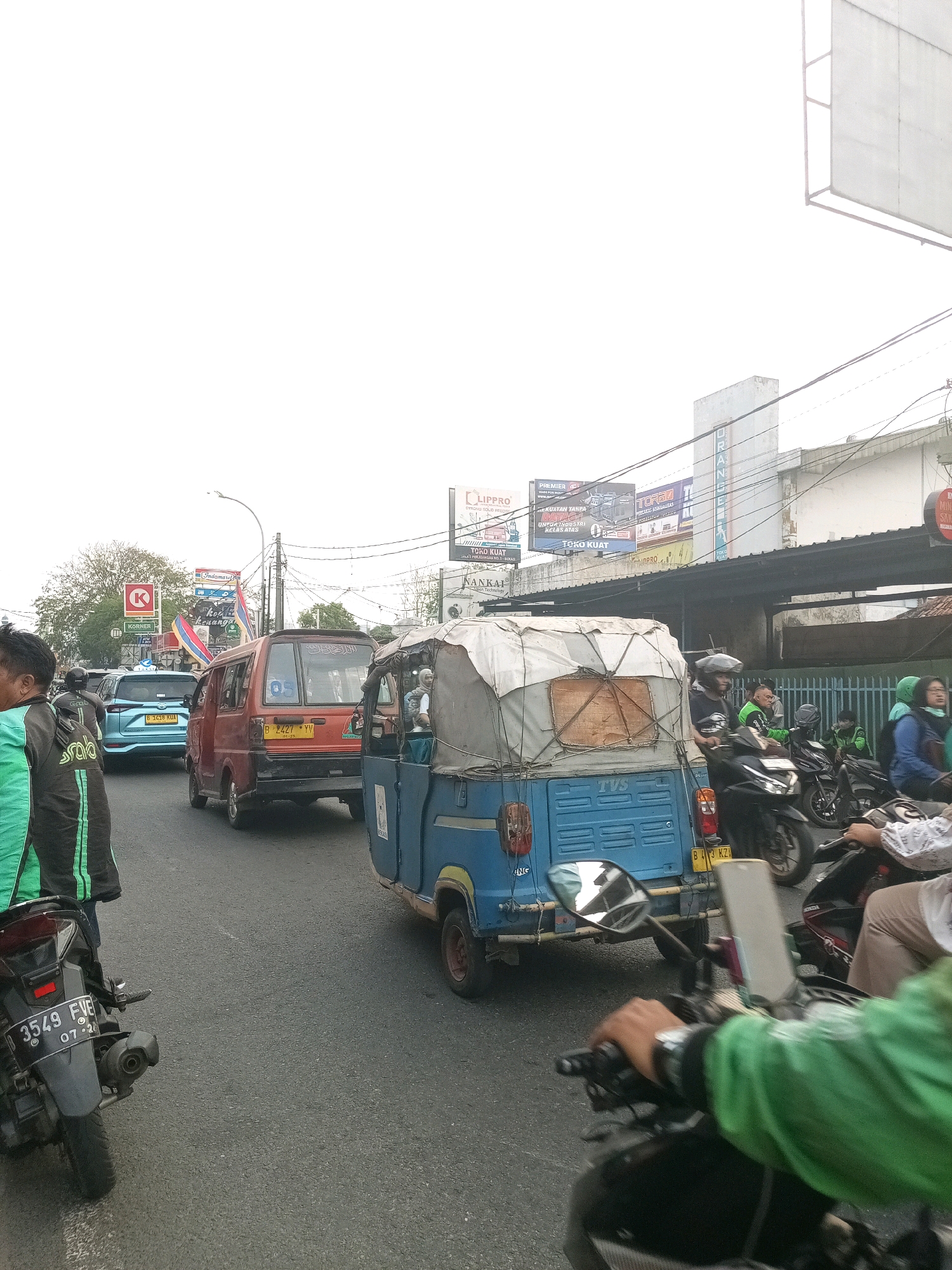 The image depicts a congested urban street scene in Bekasi, Indonesia, on an overcast day. In the foreground, a person wearing a green jacket is riding a motorcycle, their hand on the handlebars. To the left, a motorcycle with license plate "3549 FVE" is parked or stopped. Further down the road, a blue auto-rickshaw, identified with "TVS" branding, is in motion, carrying passengers. A red van with license plate "B 427 YV" is also visible, as is a light blue car with license plate "B 1418 KUA". Several other motorcycles and a larger vehicle are present in the traffic jam. Billboards and advertisements for brands like "Indomaret", "LIPPRO", and "TOKO KUAT" are displayed on buildings lining the street. The environment is typical of a busy city in Indonesia, with dense traffic and commercial signage.