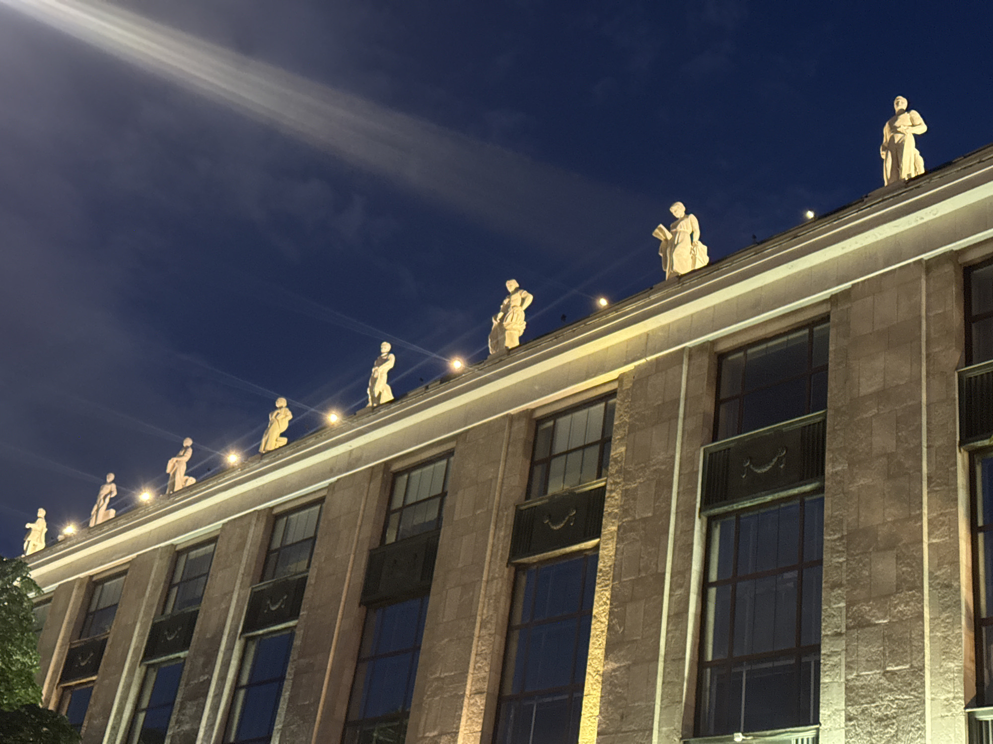 The image shows the exterior of the Moscow State University Main Building, a Stalinist Empire style skyscraper located on Sparrow Hills in Moscow, Russia. The facade of the building is made of light-colored stone blocks. Along the roofline, there are multiple statues of figures in classical attire, lit from below by warm yellow lights. The sky above is a deep blue, with a bright beam of light, possibly from a searchlight or spotlight, angled across it. The windows of the building are dark, reflecting the night sky. The overall impression is one of grandeur and monumental architecture under a night sky.