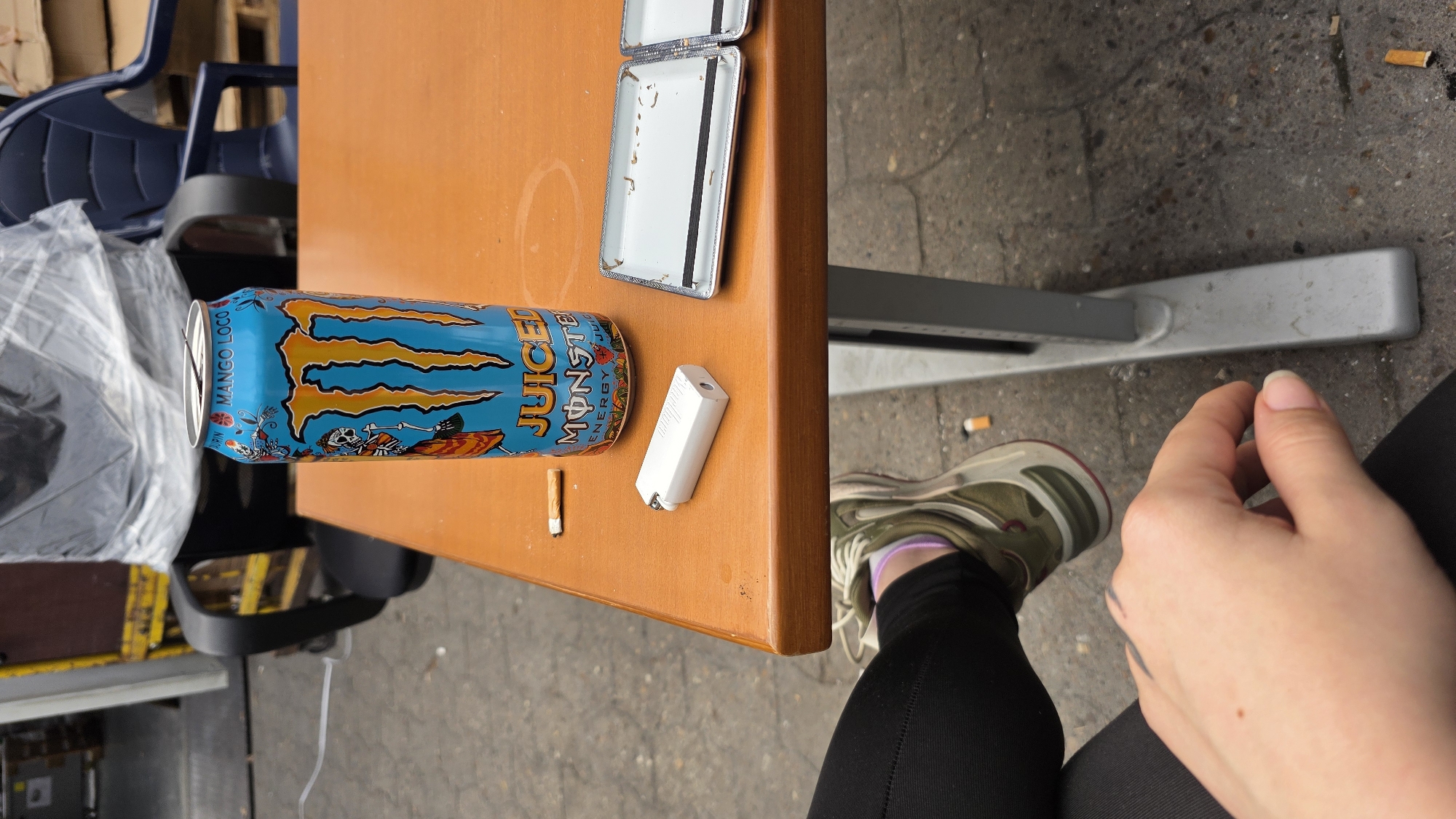 The image shows a table in an outdoor setting, likely in Achim, Germany given the context. On the table are a can of "Juiced Monster Mango Loco" energy drink, an open metal cigarette case, a white electronic cigarette, and a cigarette butt. A person is seated at the table, visible from the knees down, wearing black leggings and green and grey sneakers with purple socks. The person's hand is visible. Scattered cigarette butts are visible on the ground nearby. The table is set on a sidewalk with a metal base. A blue plastic chair and pallet are visible in the background.