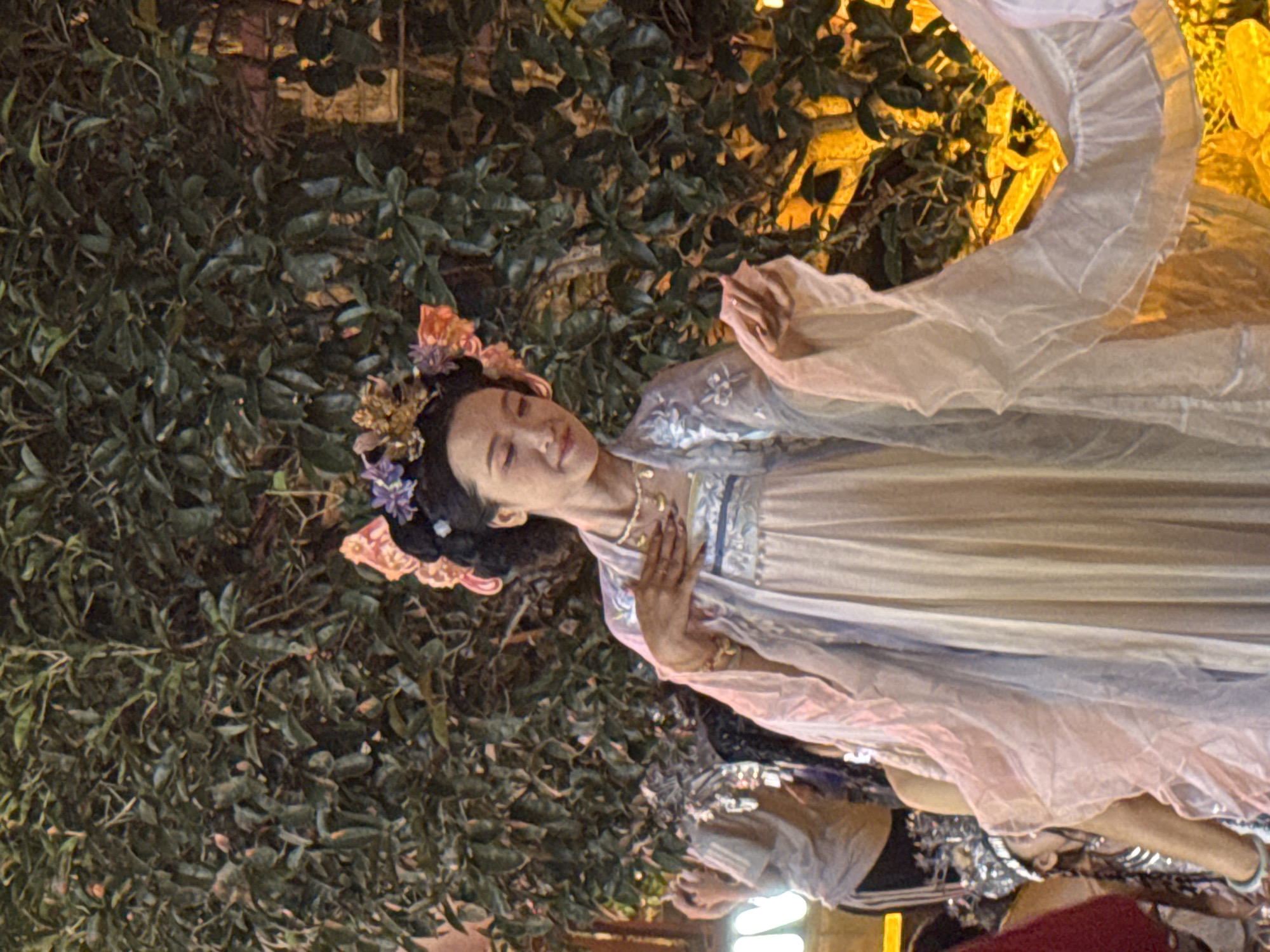 A woman in traditional Chinese attire with floral headwear is posing in front of a lush green bush. Her dress is light blue with intricate embroidery, and she wears a delicate necklace. Her right hand is on her chest, while her left hand is extended. The background is a densely packed bush with some areas illuminated by yellow light, suggesting it might be evening. Other individuals in festive attire, including a child with a bejeweled headpiece, are partially visible to the left and right of the main subject, indicating a cultural event or performance, possibly in Yuzhong District, China, given the context.