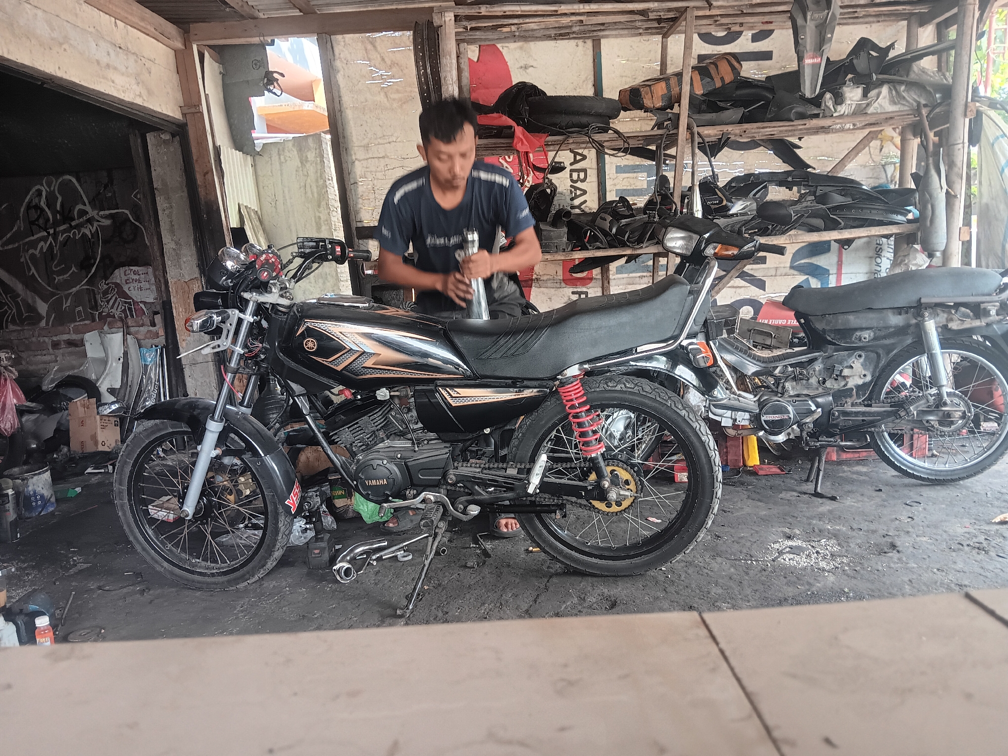 In Batursari, Indonesia, a man is working on a black Yamaha motorcycle in a dimly lit garage. The man, wearing a dark blue t-shirt, is focused on a silver cylinder he is holding. The motorcycle is parked on a dirt floor, surrounded by tools and other motorcycle parts. Another motorcycle, a black Honda, is parked behind the Yamaha. Shelves filled with various motorcycle parts line the back wall of the garage. The environment is cluttered and appears to be a functional workshop.