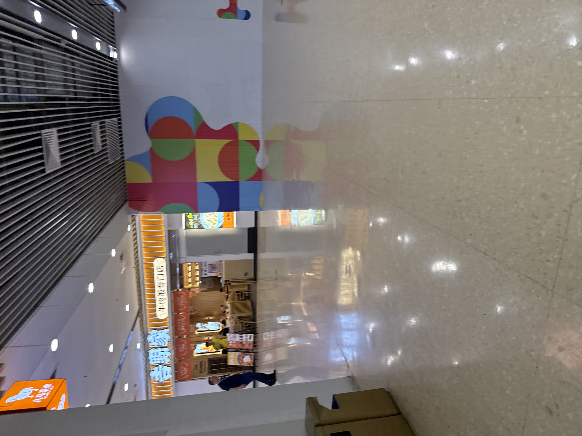 The image shows an interior view of a modern shopping mall corridor, likely in Tianhe District, China. The corridor features a polished, light-colored terrazzo floor that reflects the overhead lighting and ceiling elements. To the left, a restaurant or food establishment with a brightly lit orange fascia is visible. Signs with Chinese characters are displayed, one of which appears to be for a "Beef Noodles Specialty Store." A man in a dark suit and another individual in a yellow shirt are present near the entrance of the establishment. Further down the corridor, to the right, a large white wall is adorned with a colorful abstract geometric mural. The ceiling is a typical mall design, with metal slats and recessed lighting. A trash receptacle is located in the lower left corner of the image.
