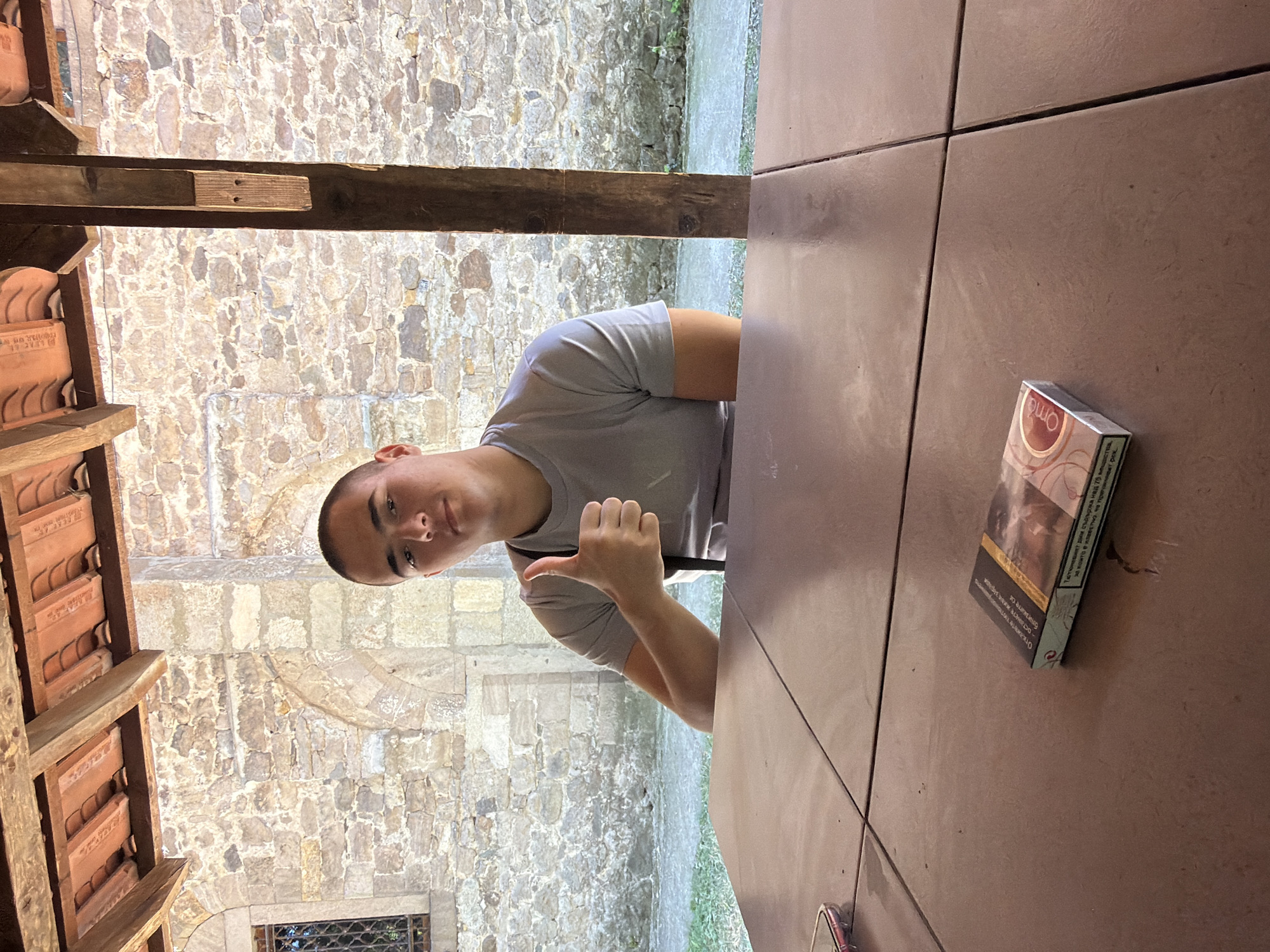 A young male is seated at a table outdoors, giving a thumbs-up gesture. He is wearing a grey t-shirt. In front of him on the table is a pack of cigarettes. The background features a stone wall and a wooden overhang. The scene appears to be in the village of Gorski Izvor, Bulgaria.