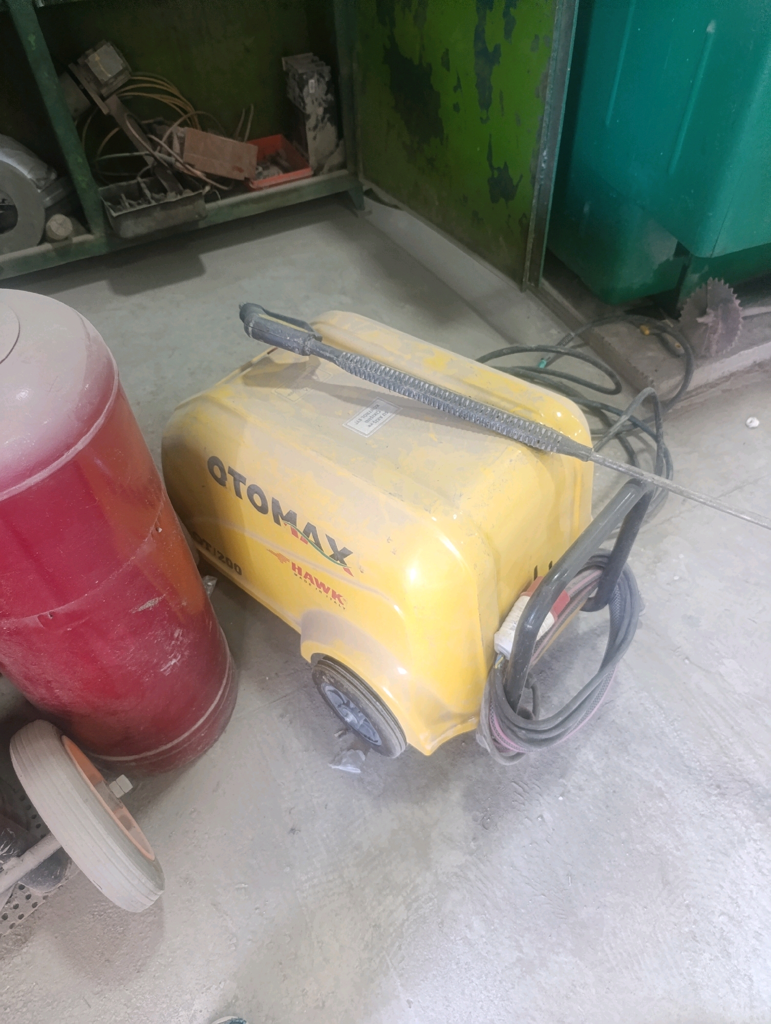 In Navoiy City, Uzbekistan, a dusty, yellow high-pressure washer, branded "OTOMAX," is positioned on a concrete floor. A red cylindrical tank sits to its left, with one of its wheels visible. The high-pressure washer has a black nozzle attached to a hose, which is coiled on the ground. The background features a green metal storage cabinet and a large green container. The scene appears to be an indoor workshop or storage area, indicated by the presence of tools and equipment within the cabinet.