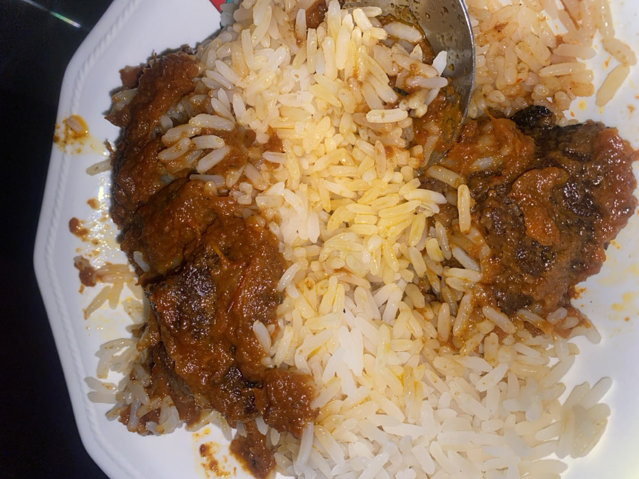 A close-up, top-down view captures a white plate with scalloped edges containing a meal, observed in Tudun Nupawa, Nigeria. The dish consists primarily of white, parboiled long-grain rice, largely covered by a reddish-brown, thick stew. Several dark, irregular pieces of meat or protein are visible within the stew, particularly concentrated towards the center and lower right. A silver-colored spoon is partially submerged into the food on the lower right side of the plate, indicating partial consumption or readiness for eating. The background is uniformly dark and out of focus, suggesting an indoor setting under low or artificial lighting conditions. No individuals are visible.