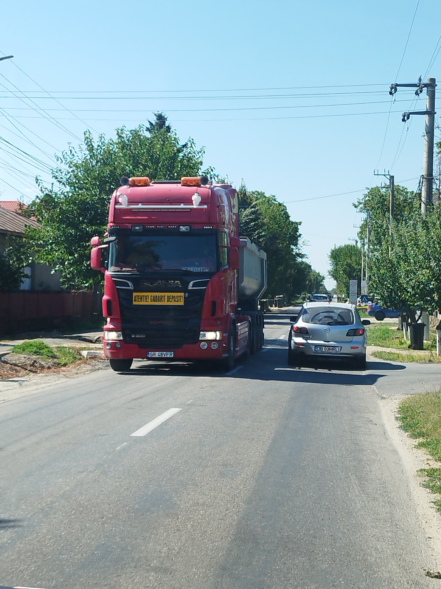 The image captures a bright, sunny day on a two-lane road in Samurcași, Romania. A large, red Scania truck, with its hazard lights illuminated, is prominently positioned in the left lane, facing the viewer. A yellow sign on the truck's front reads "ATENTIE! GABARIT DEPASIT!", indicating an oversized load. The truck's license plate is visible and reads "GR 48VFR".
To the right of the truck, a silver Mazda hatchback is in the right lane, also facing the viewer. Its license plate reads "DB 03MRL". Further down the road, other vehicles are visible, including what appears to be a white van and a dark-colored car. The roadside is lined with green trees and some residential buildings, suggesting a semi-urban or rural setting. Electrical poles and wires run along the side of the road. The road surface is asphalt with a white lane marker. The overall impression is of a typical traffic scene on a clear day.