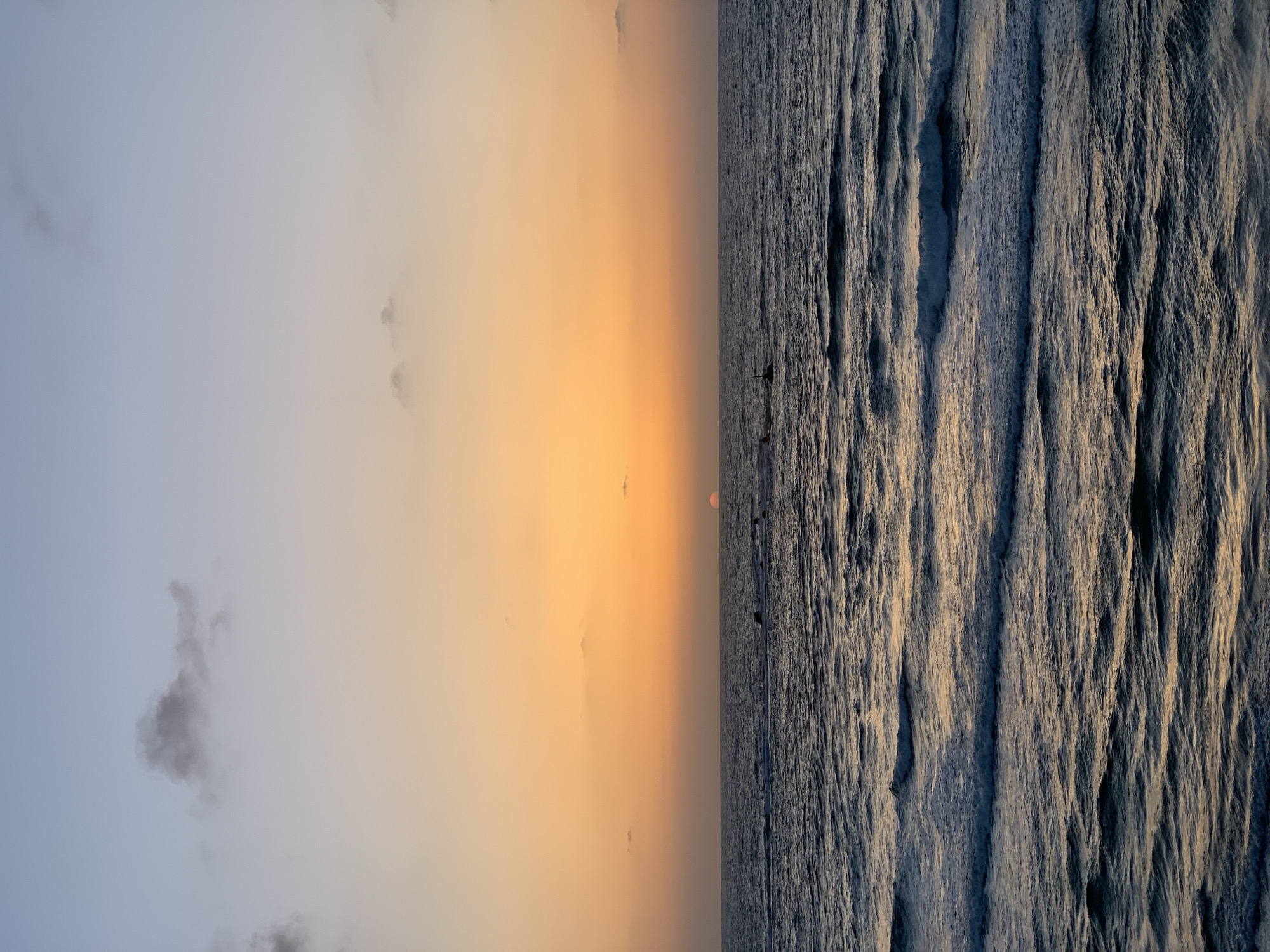 The image depicts a sunrise over the Mediterranean Sea in Tel-Aviv, Israel. The sky transitions from a pale blue on the left to a vibrant orange near the center where the sun is located just above the horizon. The sea on the right is dark and textured with waves. There are silhouettes of several people or objects visible on the water near the horizon. The overall scene is calm and serene, capturing a moment of natural beauty.