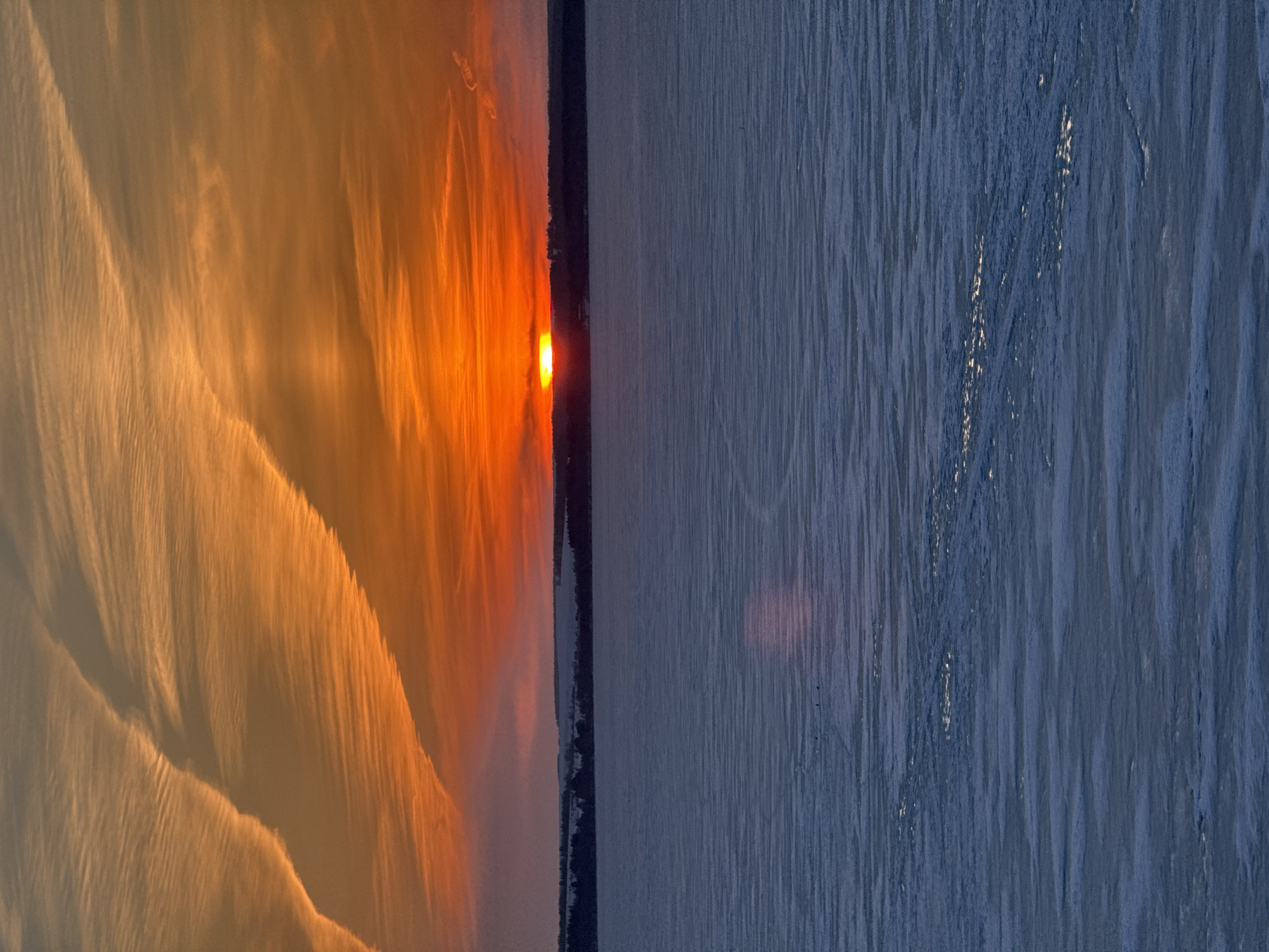 The image depicts a sunset reflected on a body of water, possibly a lake or river. The left side of the image is dominated by warm orange and yellow hues, representing the reflected sunset and possibly the clouds. On the right, the water surface appears darker with a bluish-gray tone. A narrow strip of land or a distant shoreline separates the brightly lit sky from the darker water. The presence of ice is unclear from the image. The location is stated as being in Votkinsk, Russia.