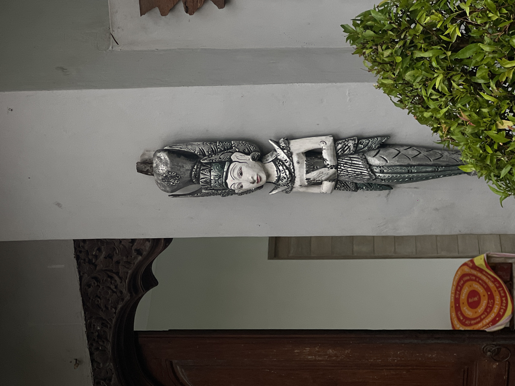 The image shows a carved wooden figure of a deity attached to a wall. The figure appears to be silver or light gray. A green bush is visible on the upper right of the figure. Underneath the figure, there is a wooden structure, possibly furniture with an ornate carved edge, and a circular object with red and yellow colors. The location is North Kuta, Indonesia.