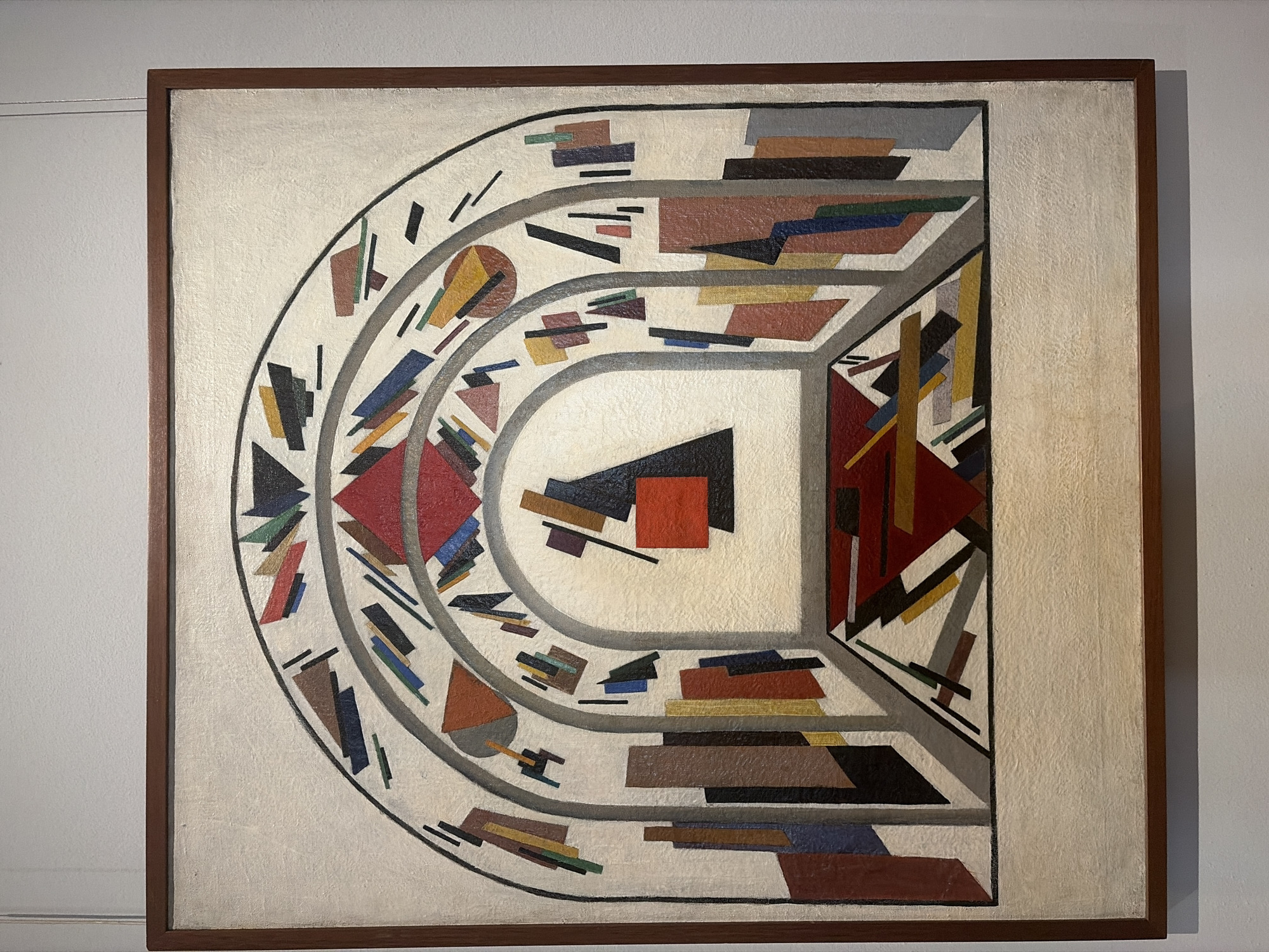 This is a detailed, forensic, matter-of-fact description of the image.
The image depicts an oil painting framed in dark wood, hanging on a plain white wall, possibly in an art gallery. The painting itself is abstract and geometric, composed of various shapes and colors.
The central motif of the painting appears to be a series of arches or tunnels, creating a sense of depth and perspective. The innermost arch encloses a central composition of geometric forms. A large black triangle with a red square inset on its left side is prominent. To the left of this triangle, there is another red square. These shapes are outlined in black and rendered with flat colors.
Surrounding the central archway are concentric bands filled with a multitude of smaller geometric shapes. These shapes are fragmented and dynamic, including triangles, rectangles, trapezoids, and lines in a palette of colors such as red, blue, yellow, brown, green, black, and gray. These elements are arranged in a way that suggests movement and energy within the arched structures.
The outer edges of the painting feature more solid, upright rectangular forms, arranged in a manner reminiscent of buildings or architectural elements. These too are rendered in muted colors like brown, gray, blue, and red. The background of the painting is a pale off-white or cream color, providing a neutral base for the geometric compositions.
The painting's style is reminiscent of Suprematism or Constructivism, art movements prominent in Russia during the early 20th century. While the image itself is a photograph of a painting, there is no visual information within the frame to indicate its specific location in Moscow, Russia, or any environmental context beyond its presentation in an art space. There are no people, actions, or interactions depicted in the painting.