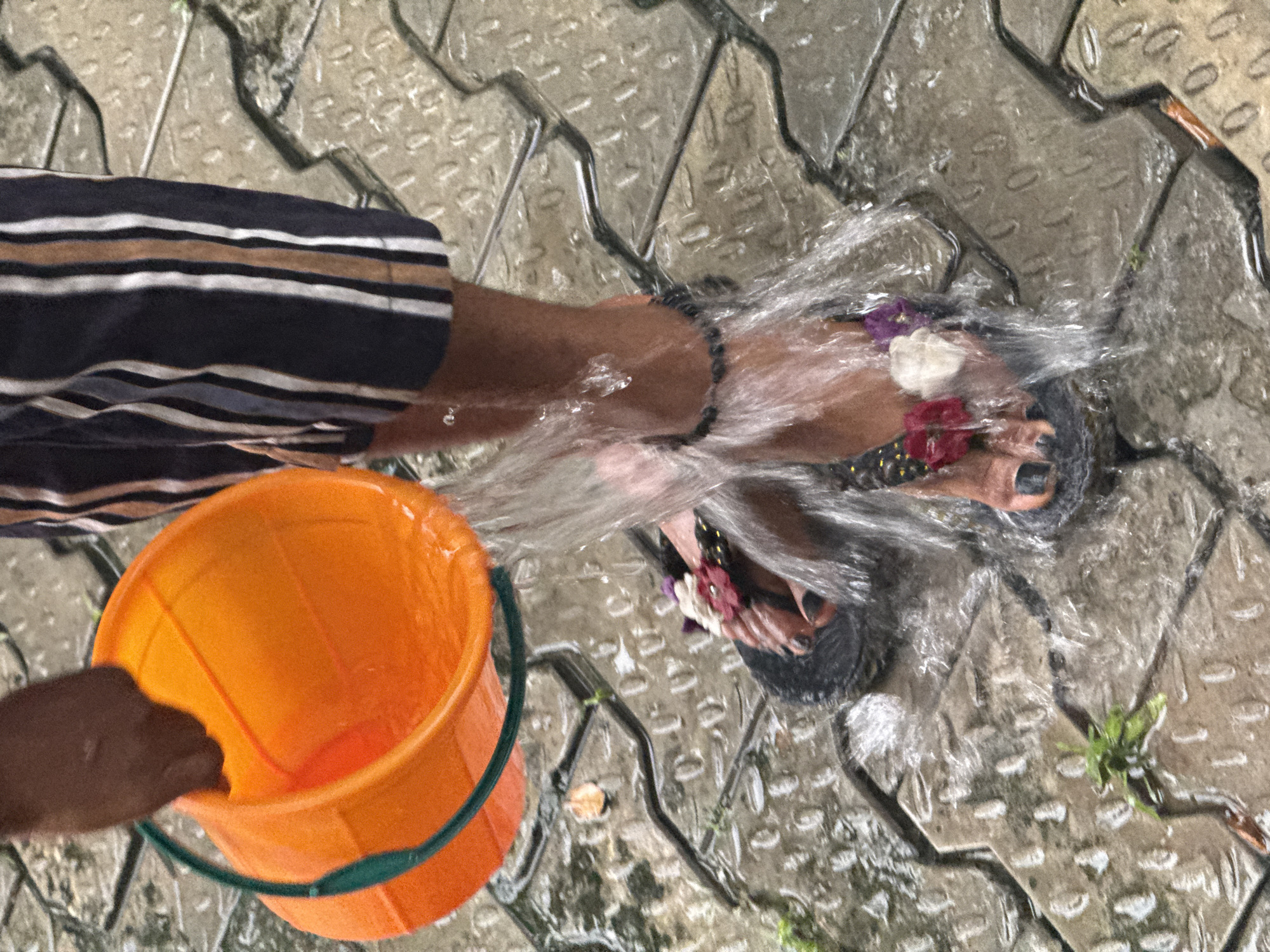 A person in striped clothing is pouring water from an orange bucket onto their feet, which are adorned with flower-decorated sandals. The scene is set on wet, patterned pavement. The location is within Ado, Nigeria.