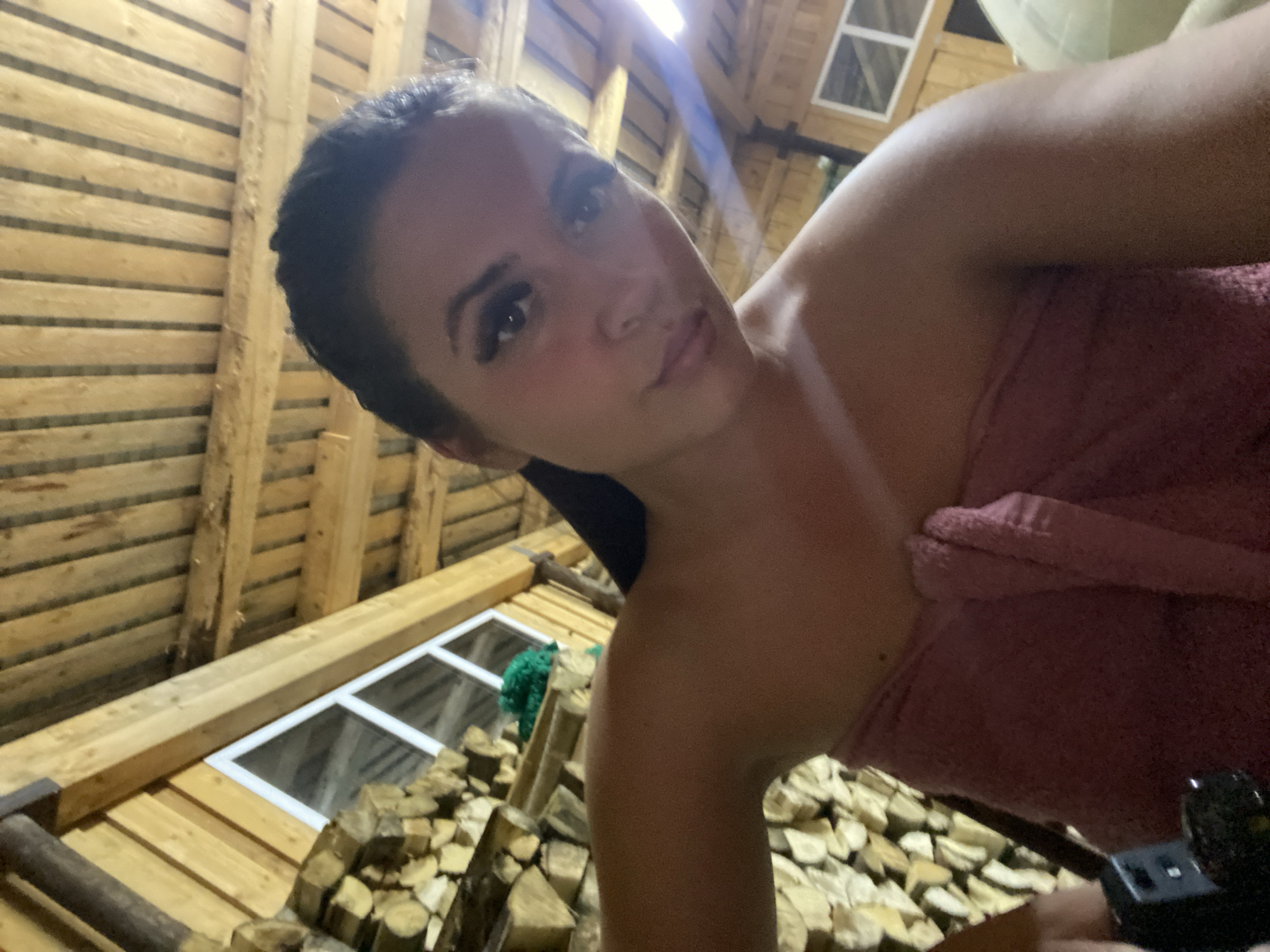 A woman is taking a selfie in what appears to be a rustic wooden structure, possibly a sauna or a bathhouse, in the city of Zhigalki, Russia. She is wrapped in a pink towel, with her hair pulled back. The background shows stacked firewood and wooden ceiling beams, suggesting a natural or rural setting.