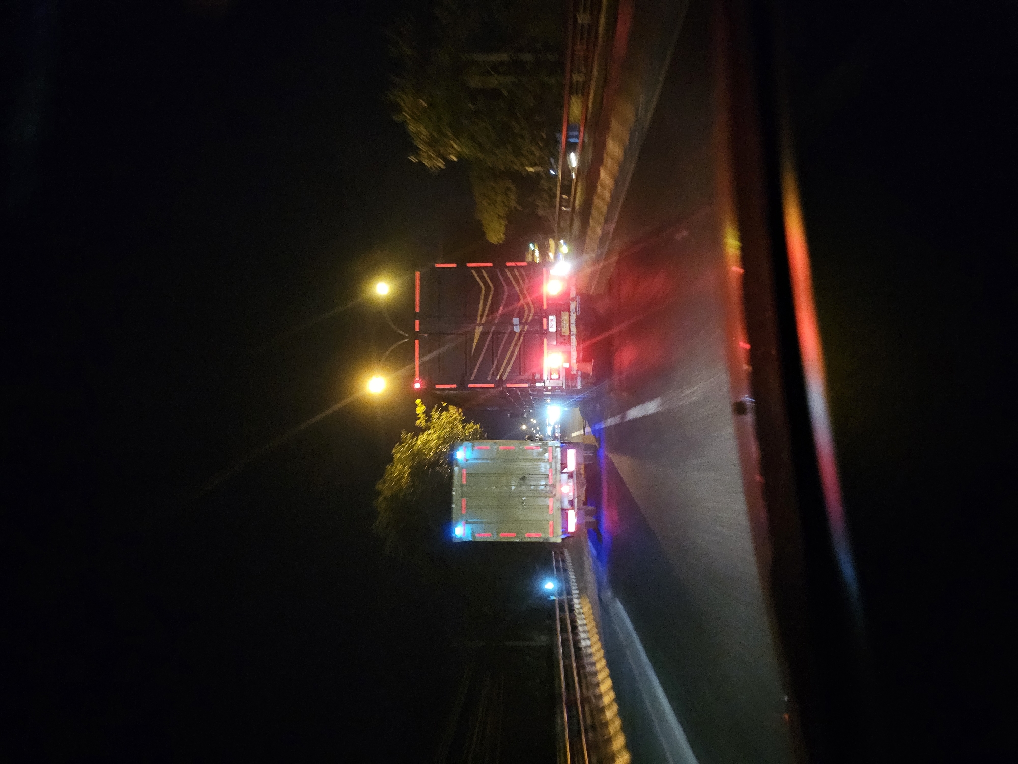 The image depicts a nighttime view from inside a vehicle on a highway, likely in Sukoanyar, Indonesia, given the context. Two large trucks are visible on the road ahead. The truck on the left has a pale green cargo box with red reflective stripes and blue lights on its upper corners. The truck on the right has a dark cargo box with red reflective stripes along its sides. Both trucks have their brake lights illuminated, casting red and pink reflections on the road surface. The highway is illuminated by streetlights and the headlights of other vehicles, creating streaks of light. Trees and foliage are visible on the sides of the road. The foreground shows the dashboard and windshield of the vehicle from which the photo was taken, with reflections of the road lights. The scene suggests travel on a highway at night, possibly during rush hour given the presence of multiple trucks.