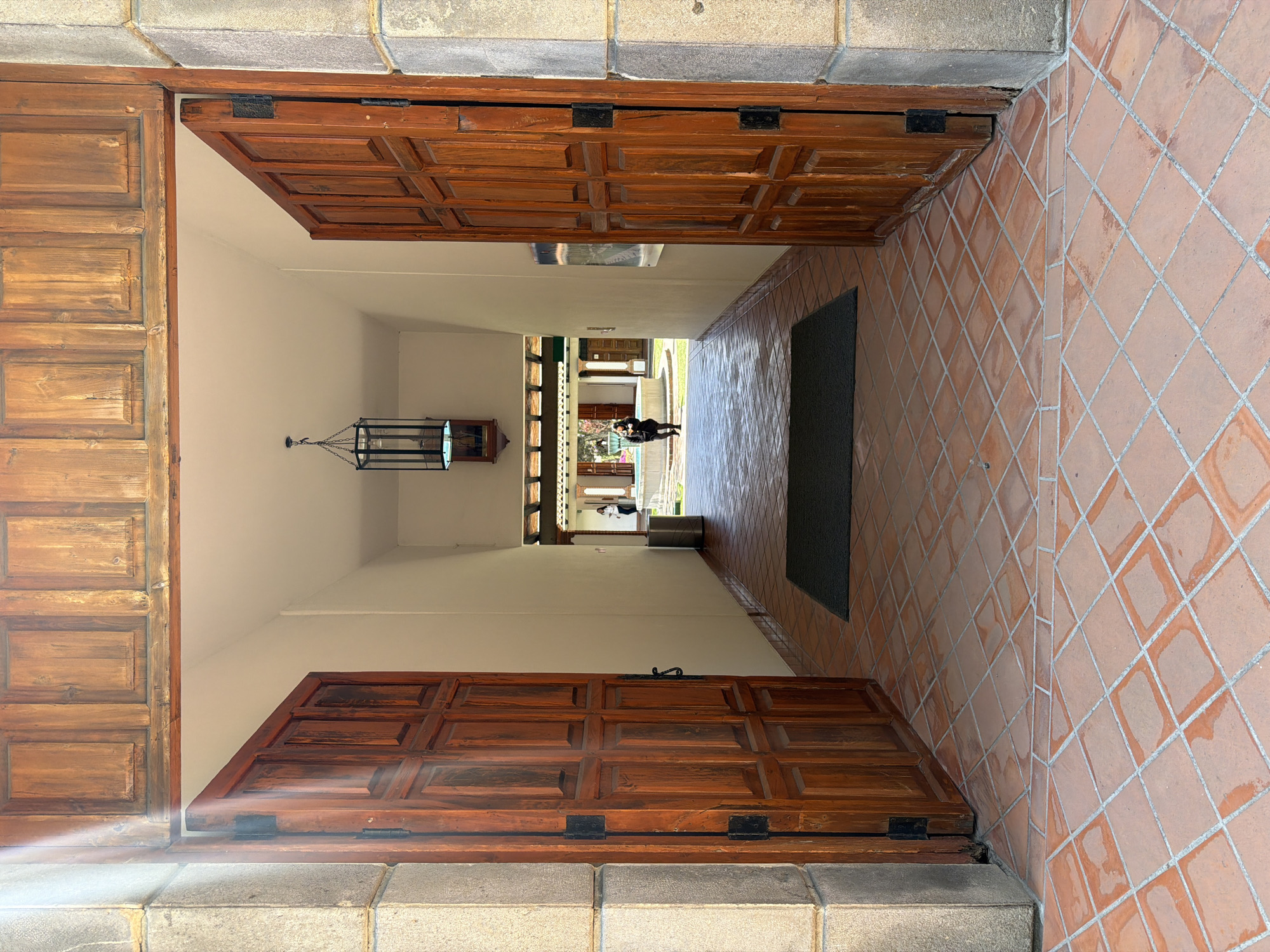 The image depicts a perspective view from an exterior vantage point, looking directly through a wide-open wooden double doorway into an indoor passageway, which subsequently leads to an open-air courtyard in San Andrés Cholula, Mexico.
The foreground is dominated by large, dark reddish-brown wooden doors, each featuring multiple recessed panels and large black hinges, fully retracted to reveal the interior. These doors are framed by rough-hewn light gray stone pillars on both sides, with a matching wooden architrave above. The floor immediately beneath the viewer and extending into the passageway is covered with terracotta-colored square tiles, laid in a diamond pattern with light gray grout lines. A dark gray rectangular doormat is positioned on these tiles, just inside the threshold.
The passageway beyond the doorway features plain, light-colored walls and a flat ceiling. A black metal lantern-style light fixture, containing a visible light source, hangs from the ceiling in the mid-section of this corridor. Below the lantern, a small wooden shelf holds an indistinct small object. Against the left wall of the passageway, a tall, cylindrical silver-toned metal trash receptacle is visible. On the right wall, a framed item, possibly a poster or map, is mounted.
The passageway opens into an sunlit outdoor courtyard area. A prominent circular, tiered fountain, constructed of light-colored stone and containing water, is visible in the center-left of the courtyard. The ground around the fountain is covered in green grass. A covered colonnade, supported by light-colored pillars and featuring dark wooden beams, spans the background of the courtyard. Two individuals are present in the courtyard: one person, dressed in dark clothing, stands near the fountain, facing away from the camera, holding an item in their hands. A second person, wearing a white top, is partially visible further back on the right side of the courtyard, near the colonnade.