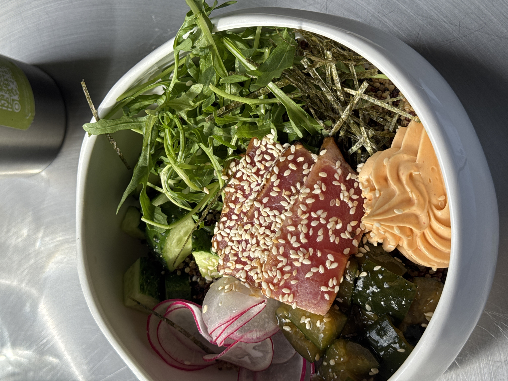 A close-up, high-angle shot depicts a white bowl filled with a variety of ingredients, likely a poke bowl or similar dish. The bowl is composed of leafy green arugula, strips of dried seaweed, slices of red radish with pink edges, diced green cucumbers, and pieces of what appears to be sesame-encrusted tuna or another type of fish. There is a dollop of orange-colored sauce, potentially spicy mayo, situated among the other ingredients. The background is a neutral, slightly reflective gray surface, which might be a table or countertop. The image appears to be taken indoors under ample lighting. There is no indication of the location being in Sochi, Russia.
