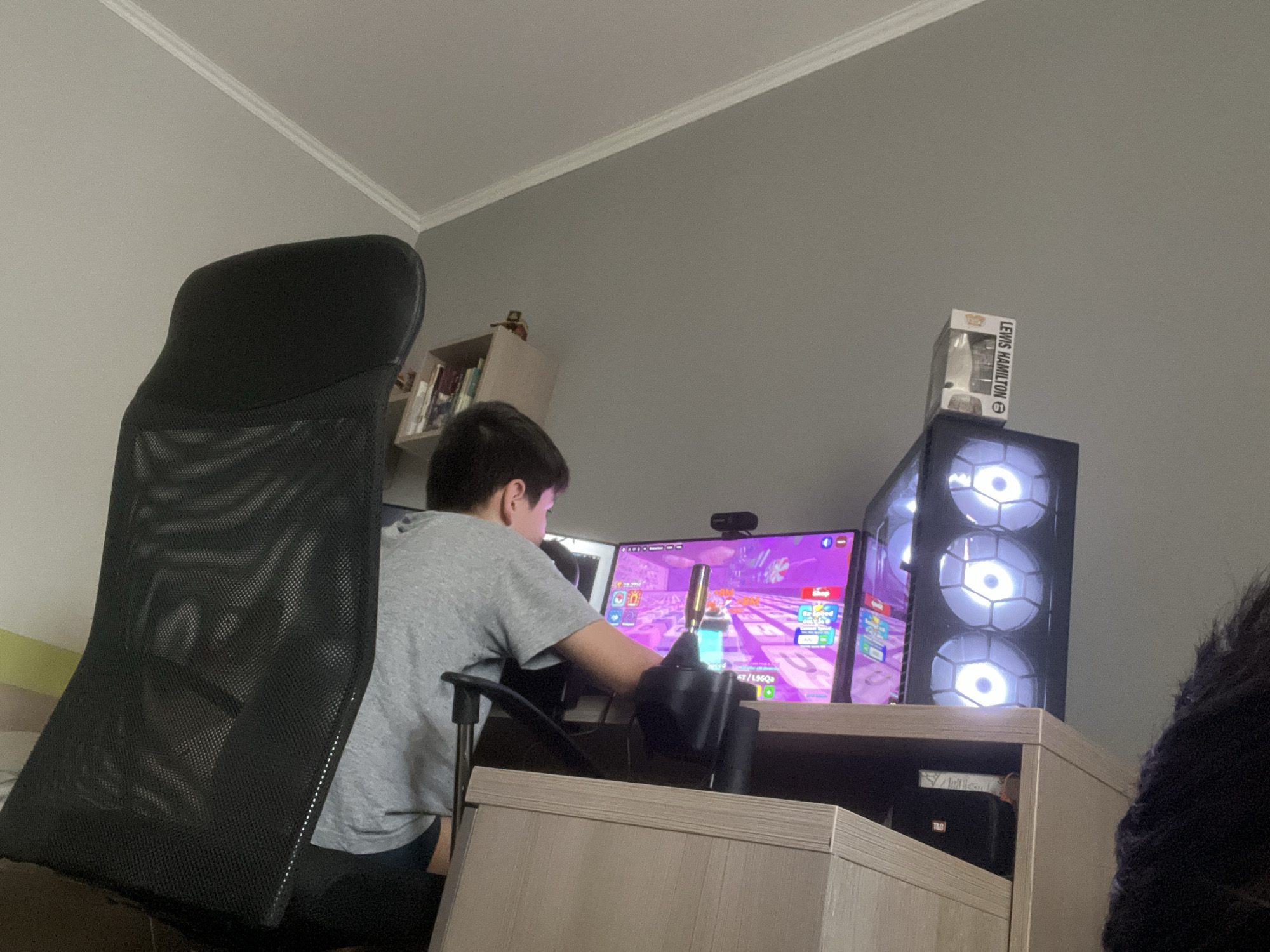 The image depicts an indoor scene, likely a home or office, in Burgas, Bulgaria. A young person, seen from the side and slightly from behind, is seated in a black mesh-back ergonomic chair, facing a computer setup. The person is wearing a grey t-shirt and dark trousers. They are actively engaged with a gaming setup, holding a controller in their hands, with their gaze directed towards a brightly lit computer monitor displaying a colorful game interface.
The computer setup includes a large monitor, a black desktop tower with multiple illuminated white fans, and a webcam positioned on top of the monitor. On the top of the computer tower, a small boxed collectible figurine is visible, with text that appears to read "LEWIS HAMILTON".
To the left of the person, the back of the ergonomic chair is prominent, showing its mesh material. To the right of the computer setup, a shelf unit is partially visible, containing books. The walls are painted a neutral color, and a white crown molding runs along the ceiling. The overall environment suggests a dedicated gaming or computer workstation.
