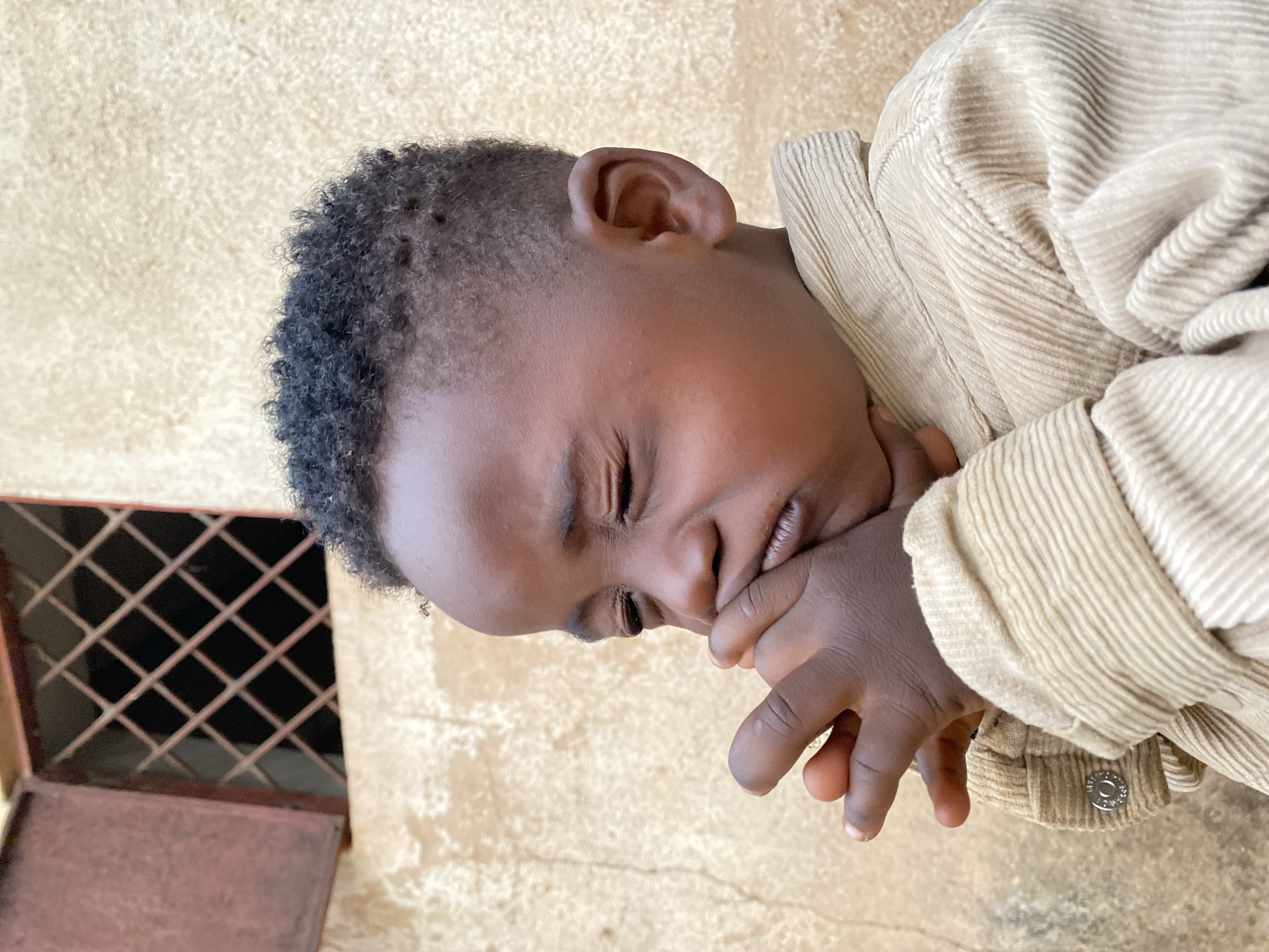 A young child with dark skin and tightly curled hair is shown from a high-angle, close-up perspective. The child is wearing a tan-colored, corduroy jacket. The child’s eyes are closed, and their hand is held close to their face, near their mouth. The background is a light-colored, textured wall with a small, red-brown metal window grate in the upper left corner. The location is Vom, Nigeria.