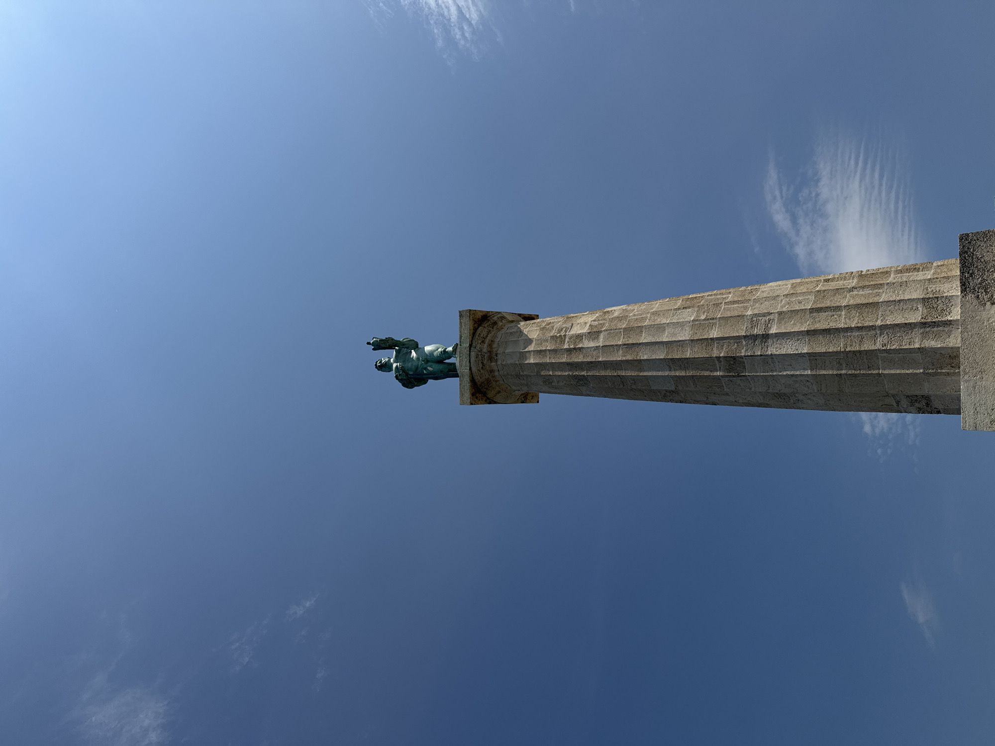A tall stone monument topped with a bronze statue stands against a clear blue sky with scattered wispy clouds. The statue depicts a male figure holding a sword and a dove. The monument's column is fluted and constructed from rough-hewn stone blocks. The perspective is from below, looking up at the monument. This is the Monument of Gratitude to France, commonly known as the Victor Monument, located in Kalemegdan Park in Belgrade, Serbia.