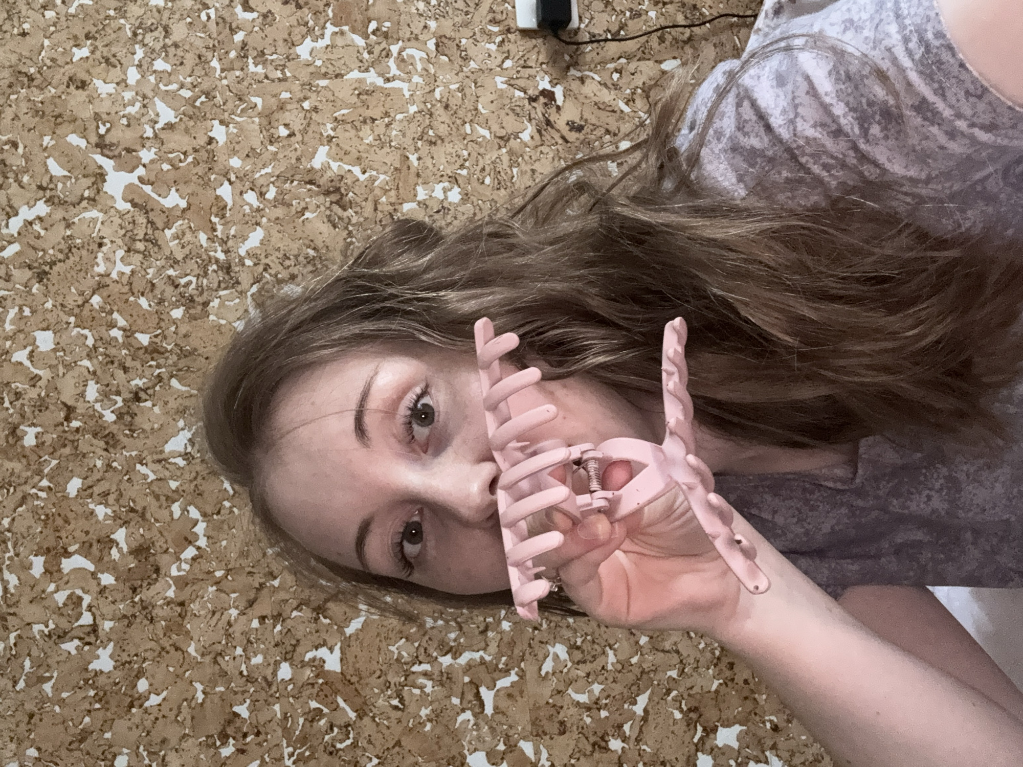 A young woman with brown hair is holding a pink claw hair clip in front of her face. She is wearing a patterned gray shirt and is lying on a corkboard surface. An electrical plug is visible at the top of the image. The location is Unknown, Unknown.
