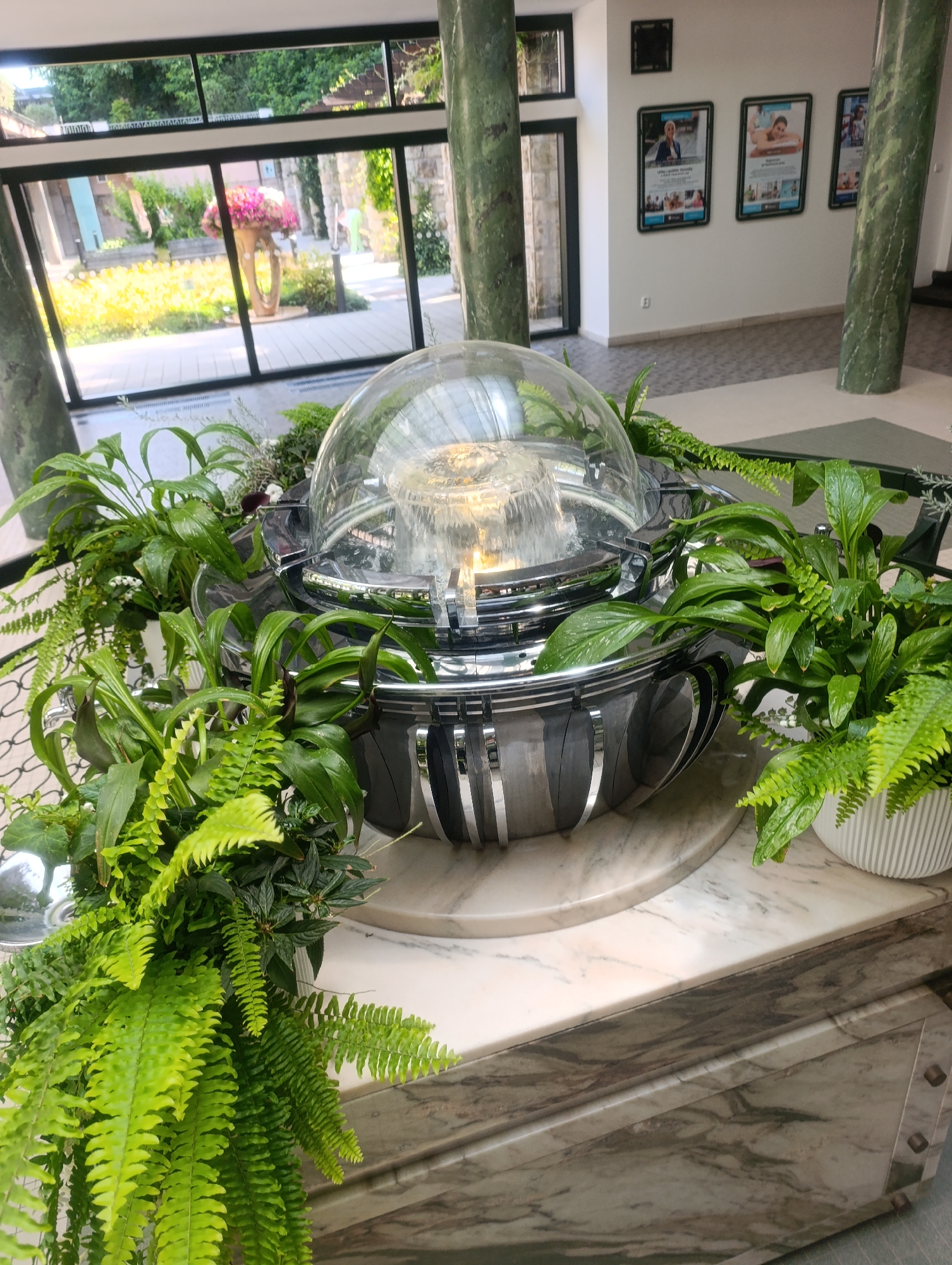 The image shows an indoor fountain with a glass dome, surrounded by lush green ferns and other plants. The fountain has a polished chrome-like base with vertical decorative elements. It sits on a marble pedestal, which is part of a larger marble structure with carved details. In the background, through large windows, a sunlit garden area is visible, featuring a large floral arrangement in a vase and greenery. The windows also reveal architectural elements of buildings and trees. The interior features tall, green marble-clad pillars and a wall with framed posters. The setting appears to be a public space, possibly a hotel lobby or a spa, given the serene ambiance and the posters which seem to advertise wellness or local attractions. The location is specified as Luhačovice, Czechia.