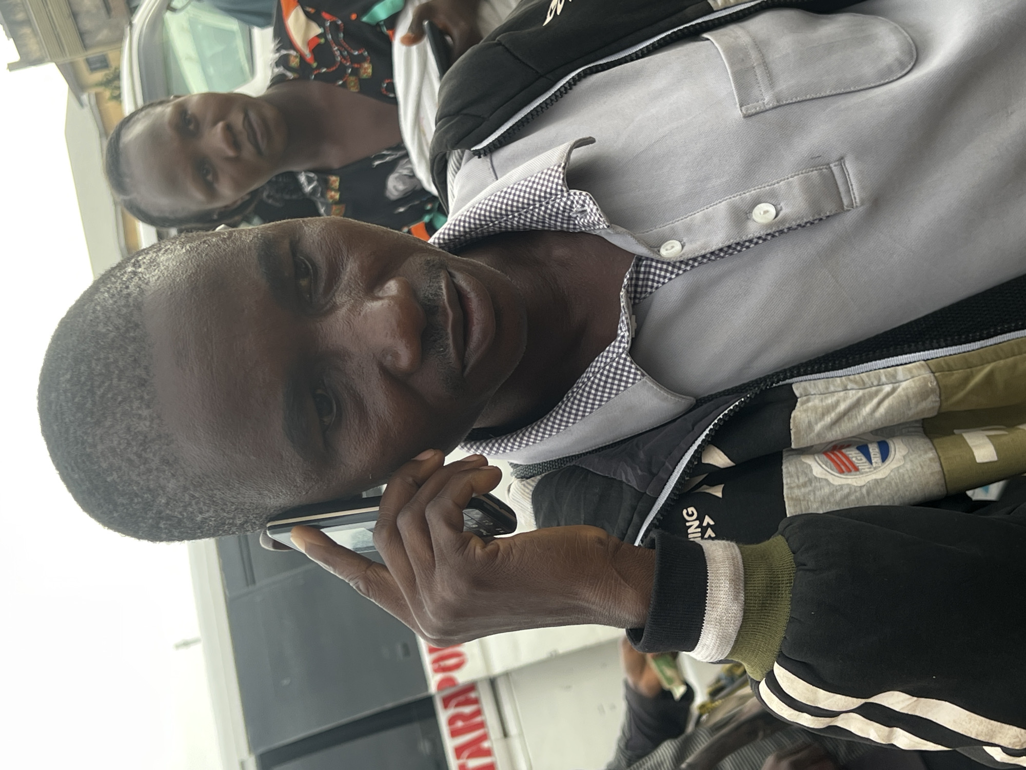 The image shows a close-up of a Black man holding a mobile phone to his right ear, appearing to be in conversation. He has short, dark hair, a thin mustache, and is wearing a grey collared shirt under a dark jacket with white and green accents on the sleeves. In the background, a Black woman with dark hair is visible, looking towards the viewer. She is wearing a patterned top with orange and black designs. A white van with text that partially reads "TARAPOLI" is behind them, suggesting a location in Jos, Nigeria. The overall environment appears to be outdoors, possibly a street or a parking area.