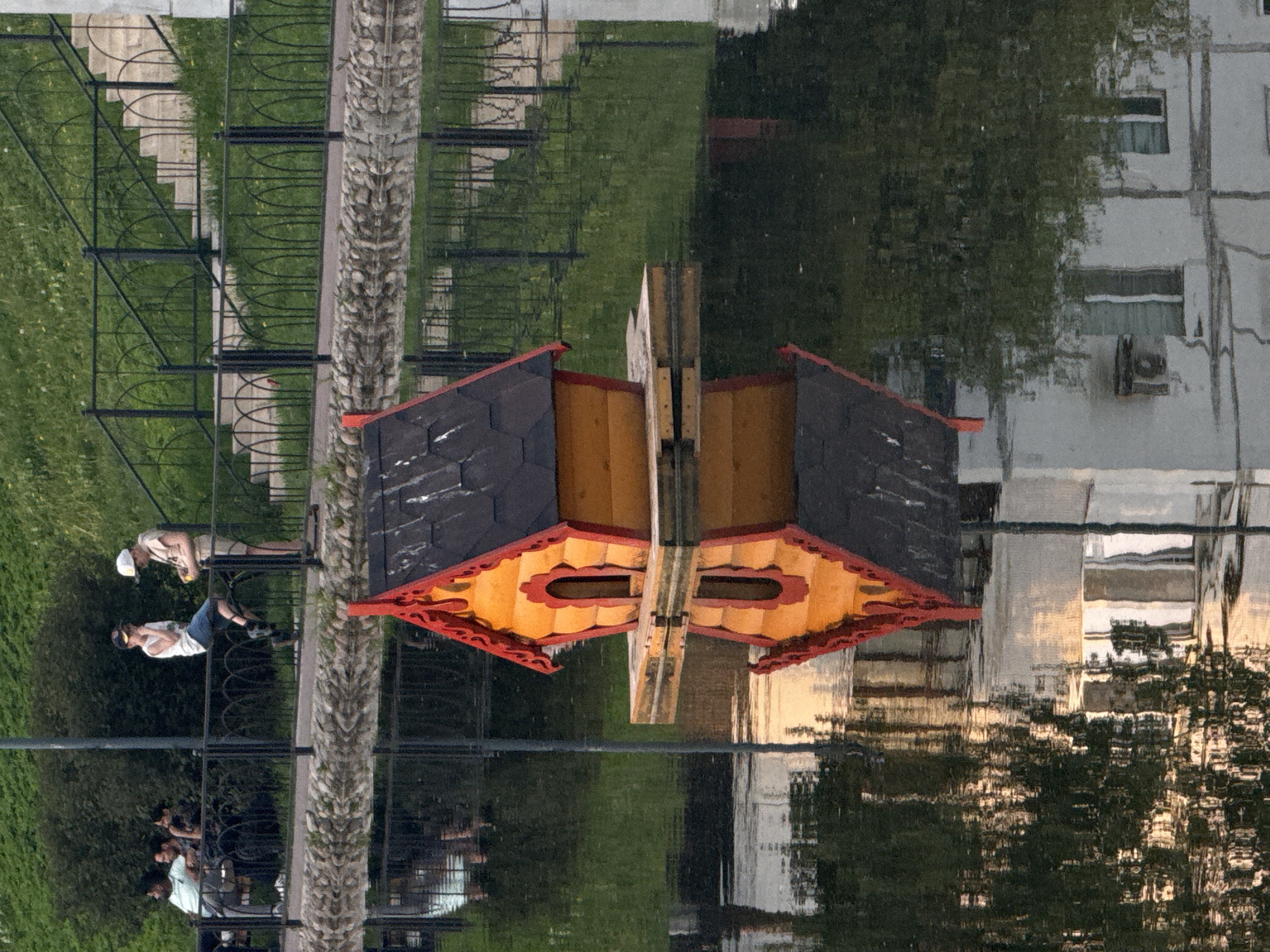 The image shows the reflection of a landscape in a body of water. A small, decorative structure with a black roof, yellow walls, and red trim is centered in the frame. A wooden post rises from the center of the structure. To the left, the reflection shows a grassy area with a black metal fence, a stone pillar, and people. Two figures stand at the fence, while others are seated nearby. To the right, the reflection reveals a building with windows and an air conditioning unit. The water surface creates a rippled effect, distorting the reflected images. The location is in the city of Moscow, Russia.