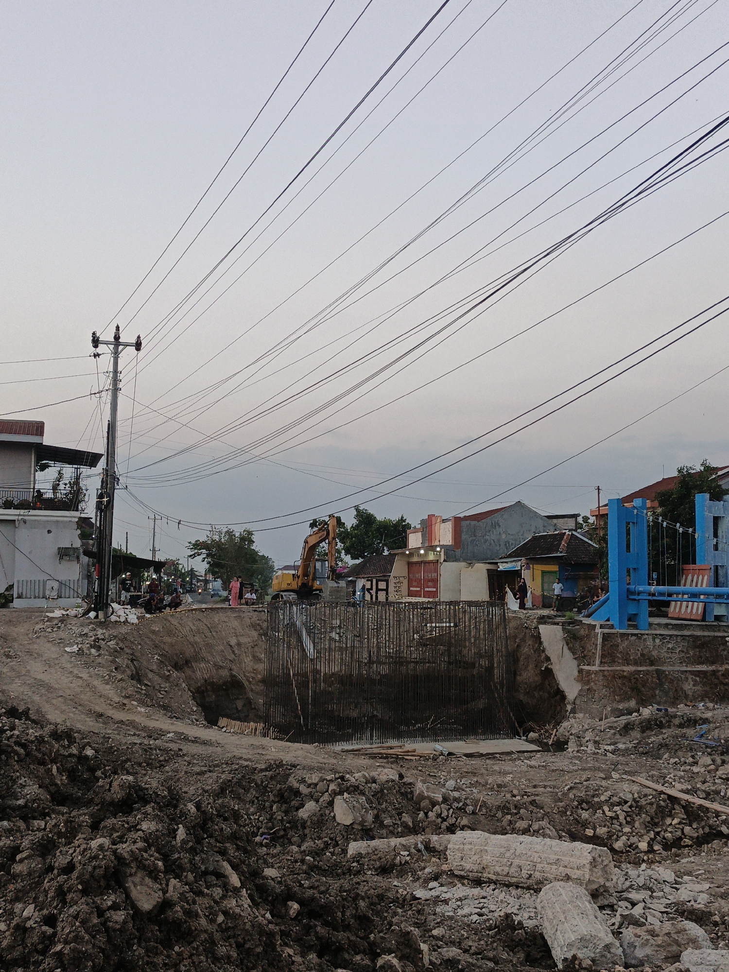 The image depicts a construction site in the city of Planggu, Indonesia, possibly near a bridge or overpass. The foreground is dominated by piles of excavated dirt and rubble, with several large concrete fragments visible. In the midground, a deep excavation is apparent, reinforced with rebar. An excavator is positioned on the edge of the excavation. Several individuals are present at the site, some appearing to be construction workers, while others seem to be observers. A power pole with numerous overhead wires stands prominently on the left side of the image, near a partially constructed building. In the background, a blue structure, potentially part of a bridge or pedestrian walkway, is visible, along with various buildings and trees, suggesting an urban or semi-urban environment. The sky is overcast.