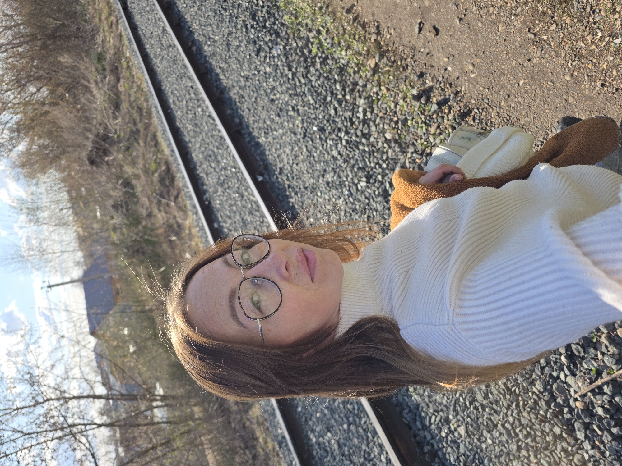 A daytime outdoor portrait features a young woman with long, straight brown hair and round, thin-framed glasses. She is looking directly at the camera with a neutral expression. She wears a white, ribbed turtleneck sweater, and a fluffy, light brown or camel-colored jacket is draped over her shoulders.
The setting appears to be next to or on a railway line. Two parallel metal railway tracks are prominent, receding into the background, with grey gravel ballast spread between and around them. The ground immediately around the tracks is covered in small stones and some sparse patches of dry grass. Beyond the tracks, the background consists of numerous bare trees and bushes, indicating either late autumn, winter, or early spring. Through the branches, patches of a blue sky with some scattered clouds are visible, suggesting clear weather. The natural light highlights her hair and glasses. No specific activity beyond posing for the photograph is depicted. There is no visible text in the image. The scene is located in Chaikovsky, Russia.