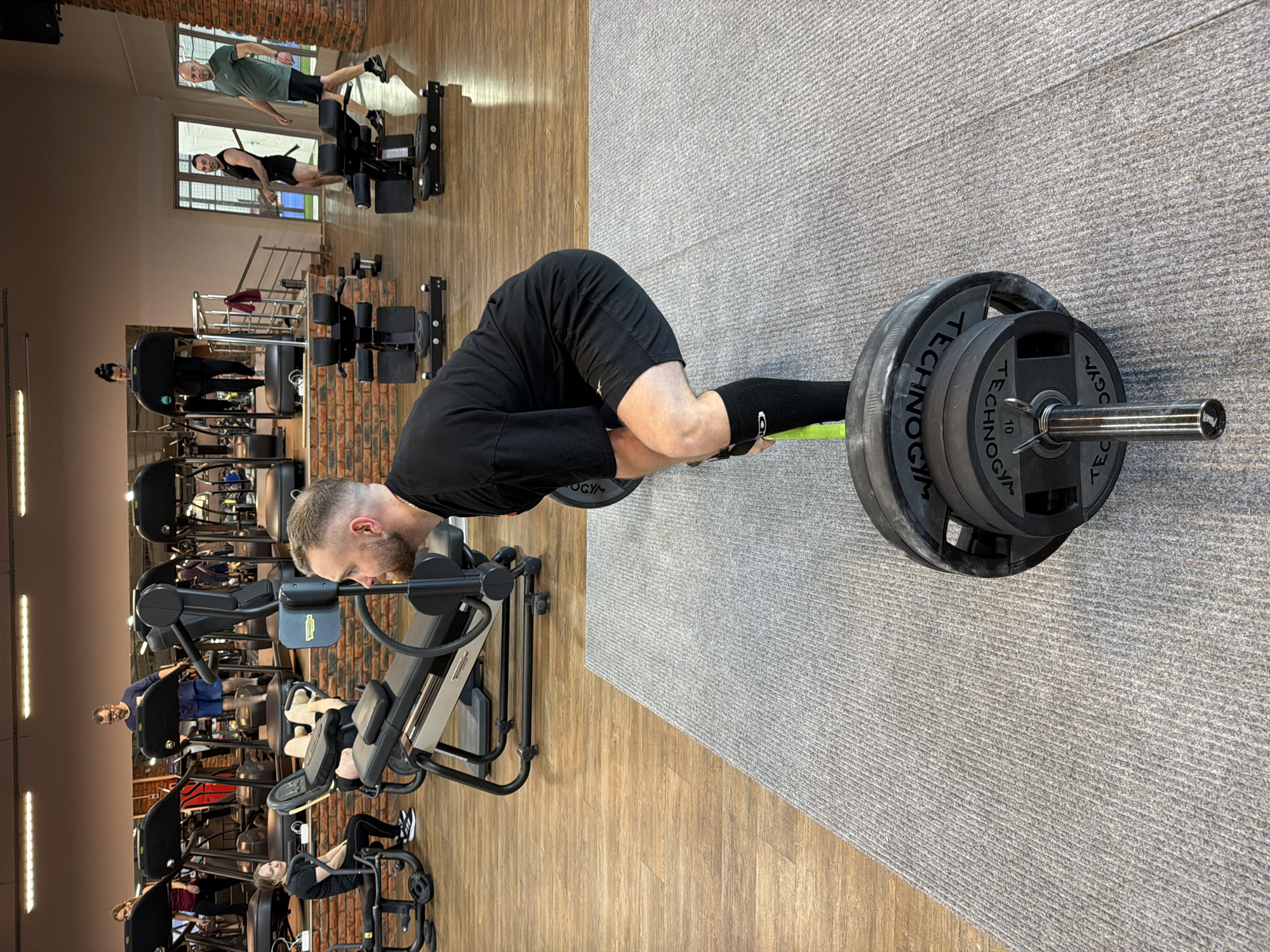 The image shows the interior of a fitness center. A man is performing a bent-over row with a barbell, loaded with several weight plates. He is wearing black athletic shorts and a black t-shirt. The barbell has black weight plates on each side, marked "TECHNOGYM" and the number "10". The floor has two sections, one with a wooden appearance and another covered in gray carpeting. Several other people are working out on various machines in the background. Location: Gym in Chaikovsky, Russia.