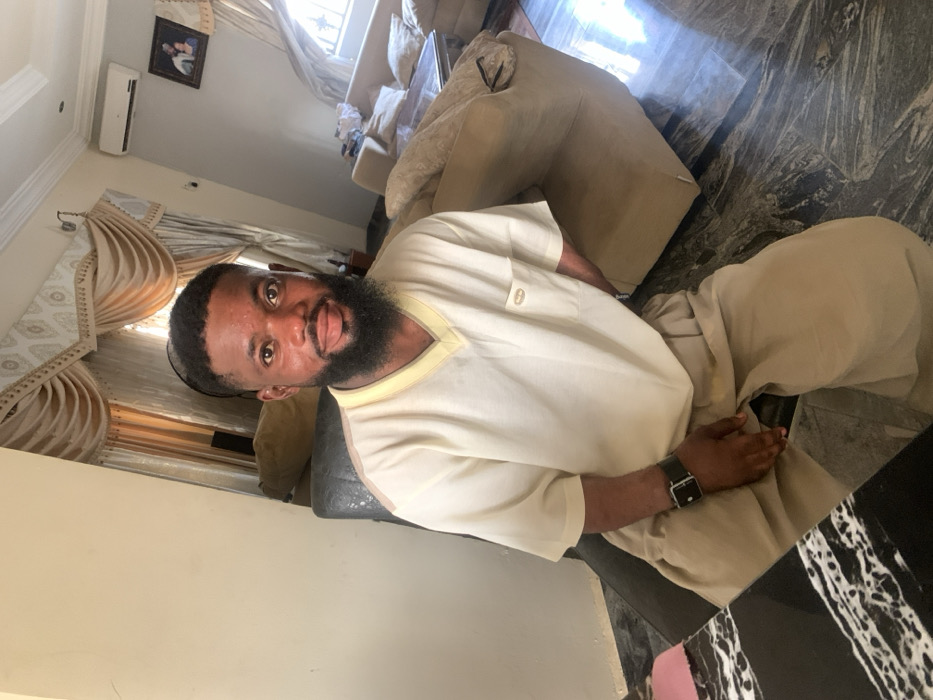The scene depicts an interior residential setting, specifically a living room, located in Eika-Adagu New Layout, Nigeria. A male subject is seated, positioned slightly off-center, looking directly at the camera with a neutral to subtly smiling expression. He possesses a dark complexion, short dark hair, and a full dark beard. The subject wears a light cream-colored short-sleeved V-neck top with a light yellow trim and a small dark emblem on the left chest pocket. He is also wearing loose-fitting light brown trousers. A black wristwatch with a square buckle is visible on his left wrist.
The subject is seated in a dark-colored chair, with a dark, white-veined marble-patterned table partially visible in the left foreground. The background reveals a room furnished with light beige upholstered sofas and a dark, reflective coffee table. An ornate window treatment featuring multiple layers of light-colored curtains with decorative valances is present. A white wall-mounted split-unit air conditioner is affixed to the upper right wall, below which hangs a framed picture. The floor is composed of dark, polished tiles with lighter patterns, reflecting ambient light. The walls are light cream in color. The room is illuminated by natural light, likely from the visible window.