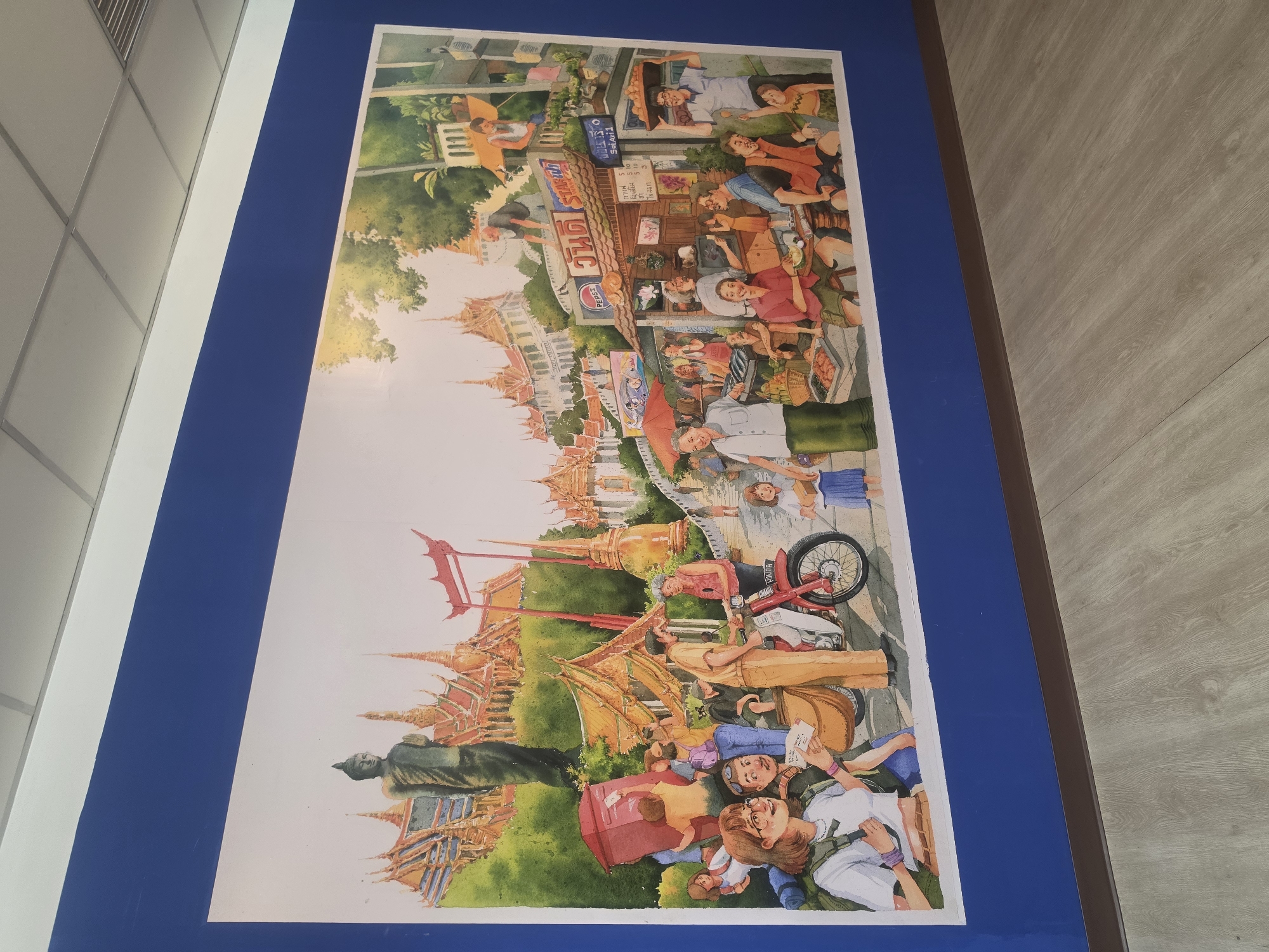 The image is a painting or print depicting a scene in Bangkok, Thailand. The artwork is mounted on a blue background and displayed indoors.
The painting includes several distinct scenes, likely depicting various tourist activities or landmarks. Prominently featured is a large Buddha statue and ornate Thai architecture, potentially representing a temple complex. In the upper part of the image, there is a depiction of a street scene with food stalls and people, suggesting a market area. Other elements visible are figures riding a motorcycle, a tall red structure (possibly a historical landmark), and a group of people lying on the ground, appearing relaxed or possibly tired from travel. Overall, the artwork seems to capture a vibrant and diverse view of Bangkok.