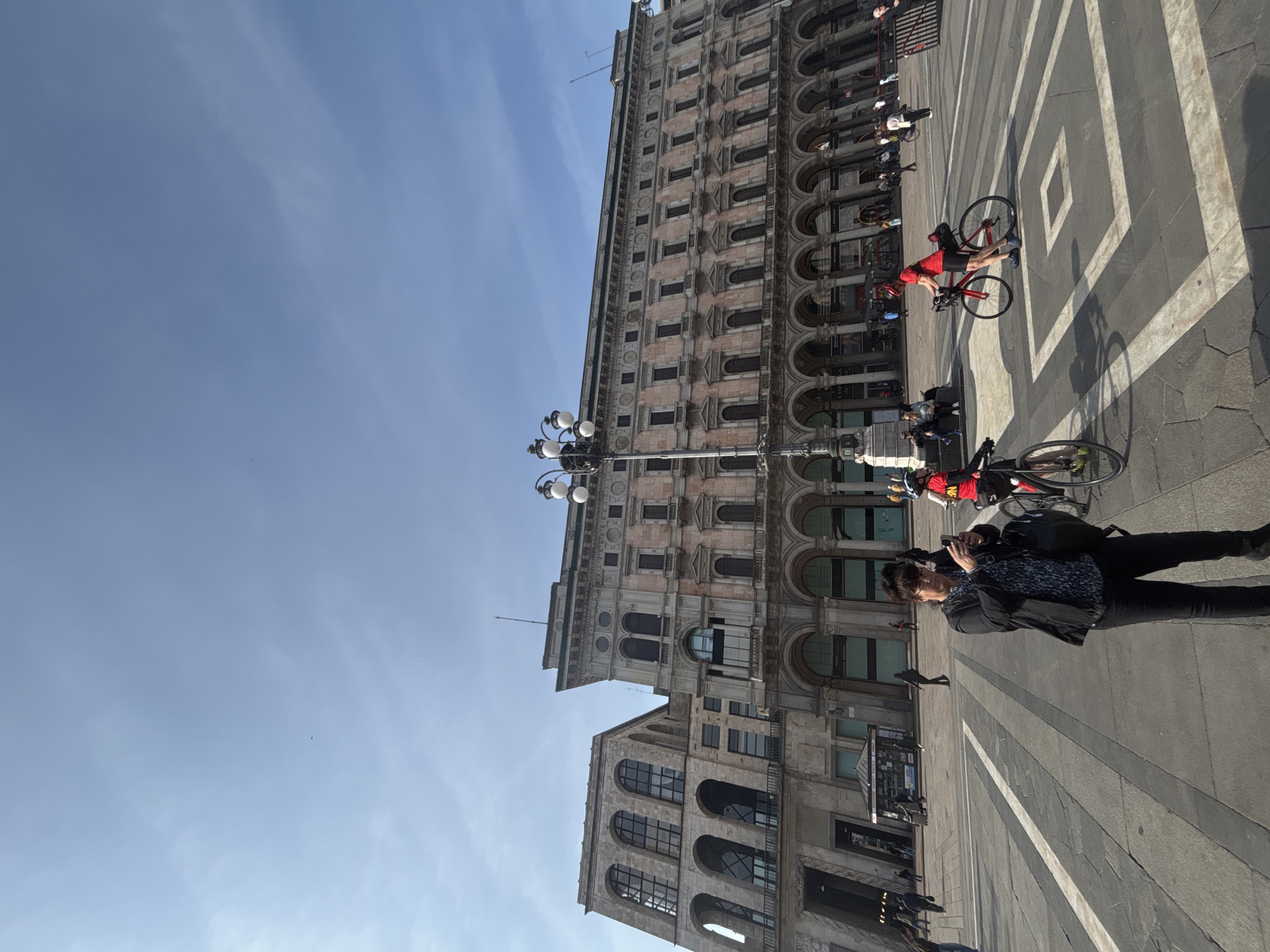 The image shows a bustling scene in the Piazza del Duomo in Milan, Italy. The Duomo's architecture is visible in the background. Two people are riding bicycles in the foreground, while a person is standing, looking at their phone. The ground is paved with a geometric pattern of light and dark stones. A lamppost is centered. Several people are walking and standing along the buildings. The sky is clear and blue.