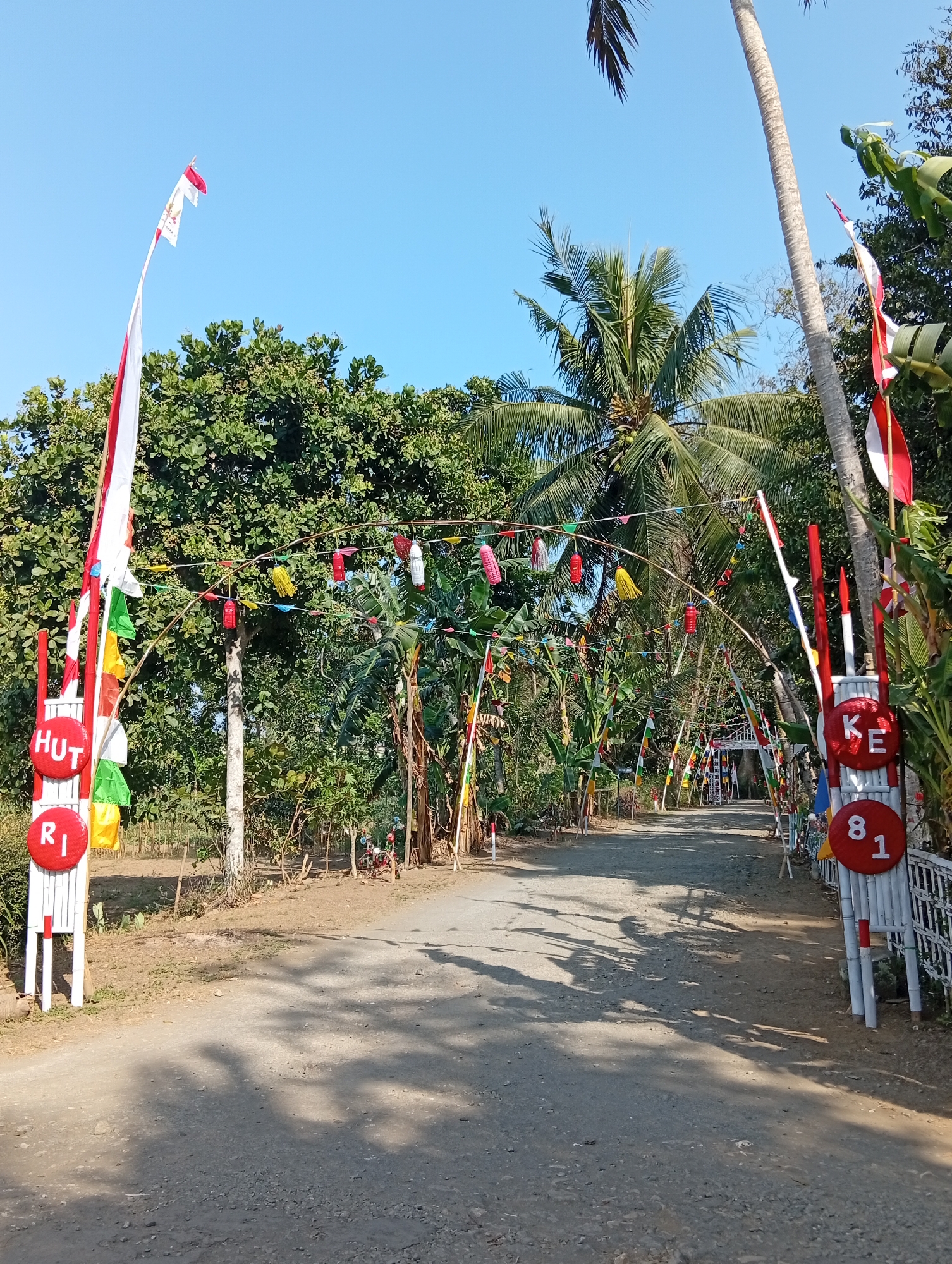 The image depicts a dirt road in Miritpetikusan, Indonesia, decorated with Indonesian flags and colorful festive banners. The road leads through a lush green environment with abundant trees, including palm trees. On the left side of the road, a white structure displays the letters "HUT" and the number "R1" in red circles. On the right side, a similar structure shows the letters "KE" and the numbers "8" and "1" in red circles. Small flags and lanterns are strung across the road, creating an archway effect. The sky is clear and blue, indicating a sunny day. The scene appears to be set up for a local celebration or event.