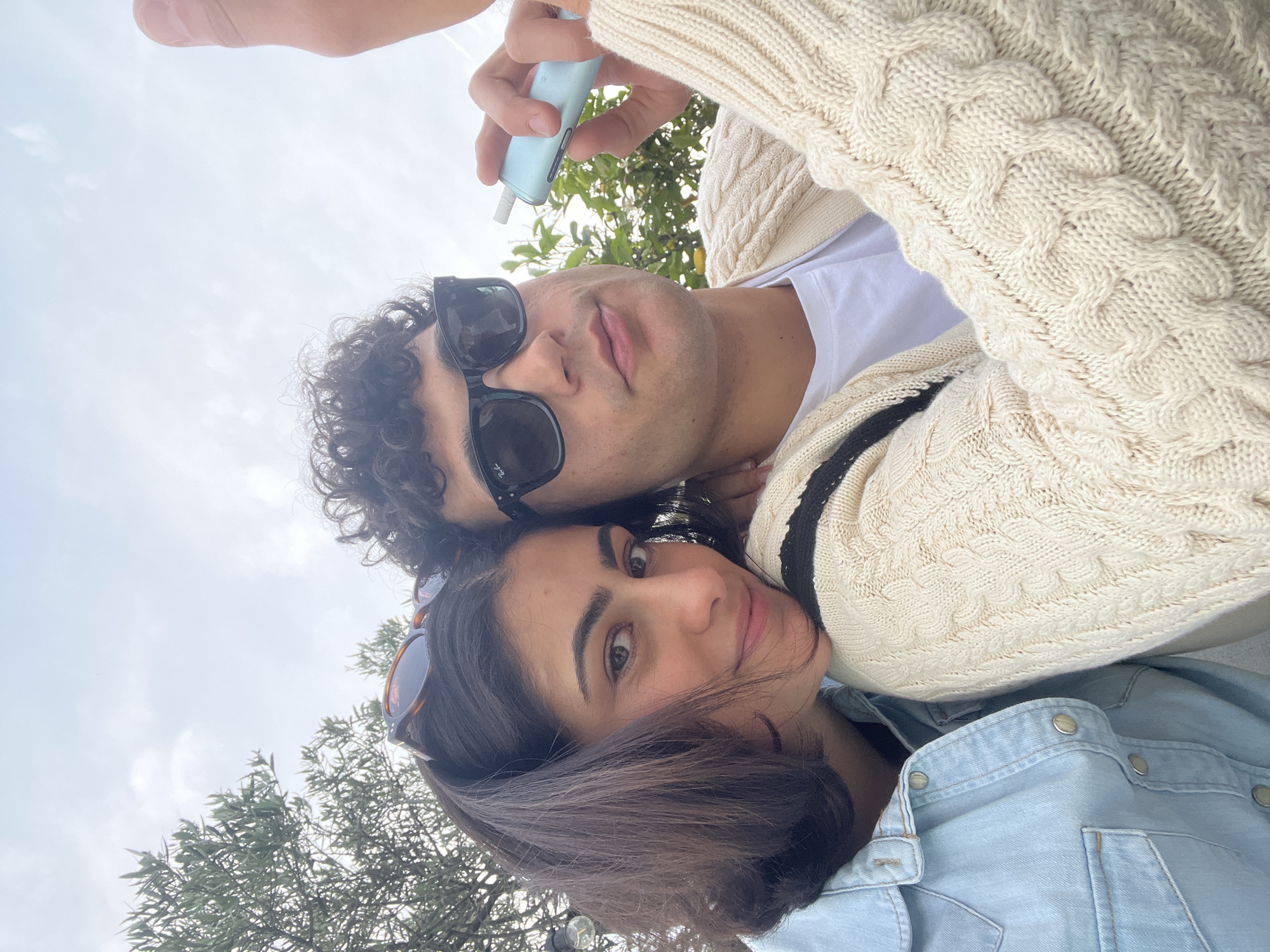 This low-angle selfie captures two young adults, a man and a woman, outdoors during the daytime. The perspective looks up towards the subjects against a light, possibly overcast, sky.
The man, positioned slightly above and behind the woman, has dark curly hair and wears black sunglasses, revealing "Ray-Ban" text on the frame. He is dressed in a white t-shirt layered under a cream-colored cable-knit cardigan featuring a black stripe near the collar. His left hand is visible at the top of the frame, holding a light blue electronic device, possibly a vape pen.
The woman is in the lower-middle part of the image, resting her head close to the man. She has dark, straight hair and appears to have sunglasses pushed up onto her head. She is wearing a light blue denim shirt or jacket and looks directly at the camera with a subtle, relaxed smile.
The background shows a bright, diffused sky, suggesting an overcast day or soft daylight. Green foliage from trees is visible, indicating an outdoor setting such as a park or garden. The overall atmosphere is casual and relaxed, capturing a shared moment in what is contextually understood to be Milan, Italy.