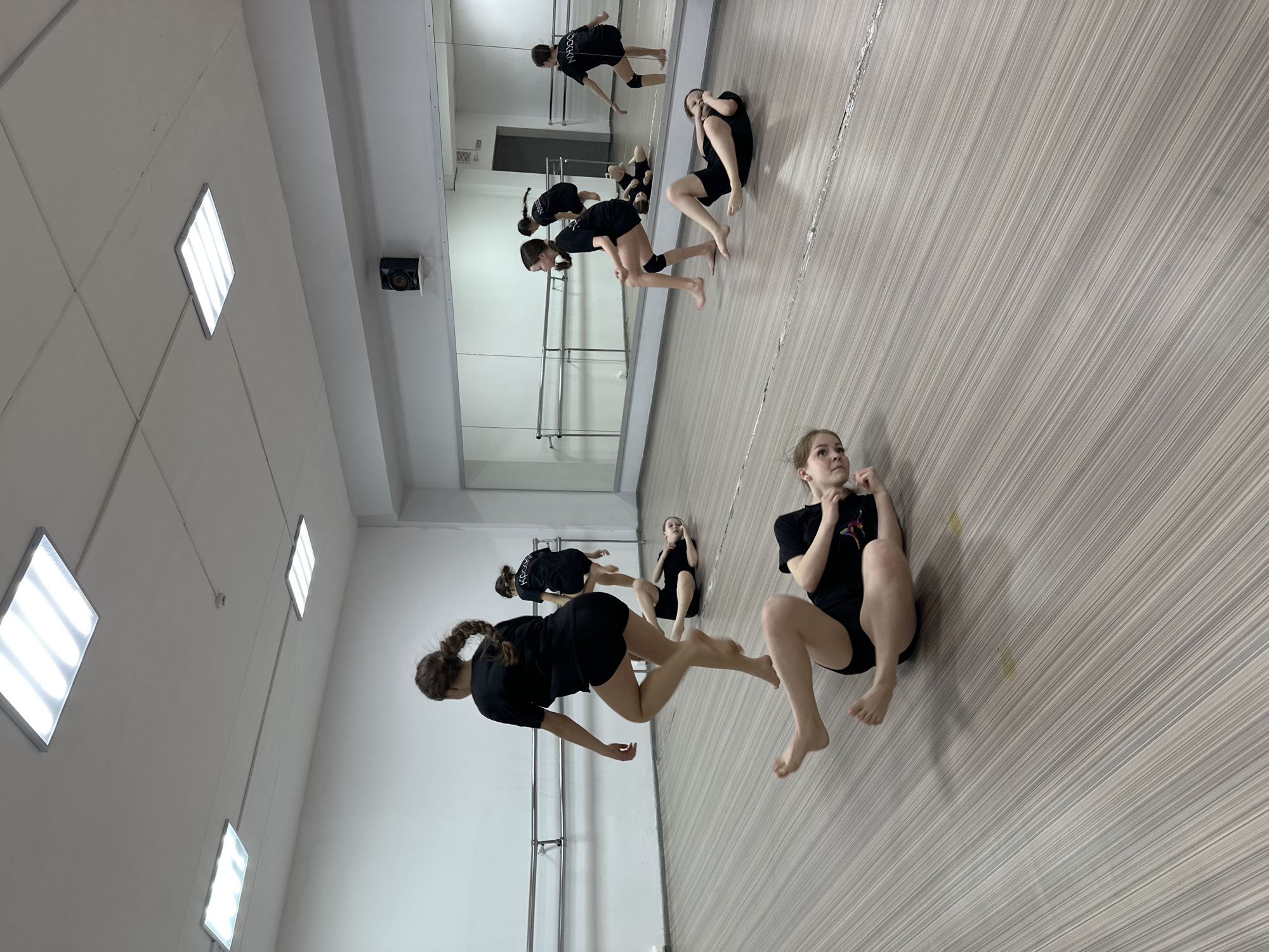 An indoor studio in Votkinsk, Russia, displays six young females engaged in a dance or exercise session. The studio features a light-toned, striped wood-look floor, plain white walls, and a large mirrored wall on the right equipped with two parallel metal barres. The white ceiling contains five rectangular LED panel lights, a small white smoke detector, and a black speaker mounted on a shelf above the mirror. All individuals wear uniform black t-shirts and black shorts; some have black knee pads, and all are barefoot. In the foreground, one female is captured mid-air, with her left foot off the ground and right leg bent, while another lies on her back with bent knees, looking towards the camera. Further back, a third female is also mid-air, rising from a squat, wearing knee pads. A fourth lies on her side with knees drawn up. Reflections in the mirror show additional participants, including one with "NADC" or "NADCC" on her shirt and another with Cyrillic characters. A portion of a fifth female's face is visible in the extreme bottom-left, looking up. The participants are in various states of dynamic movement or rest.