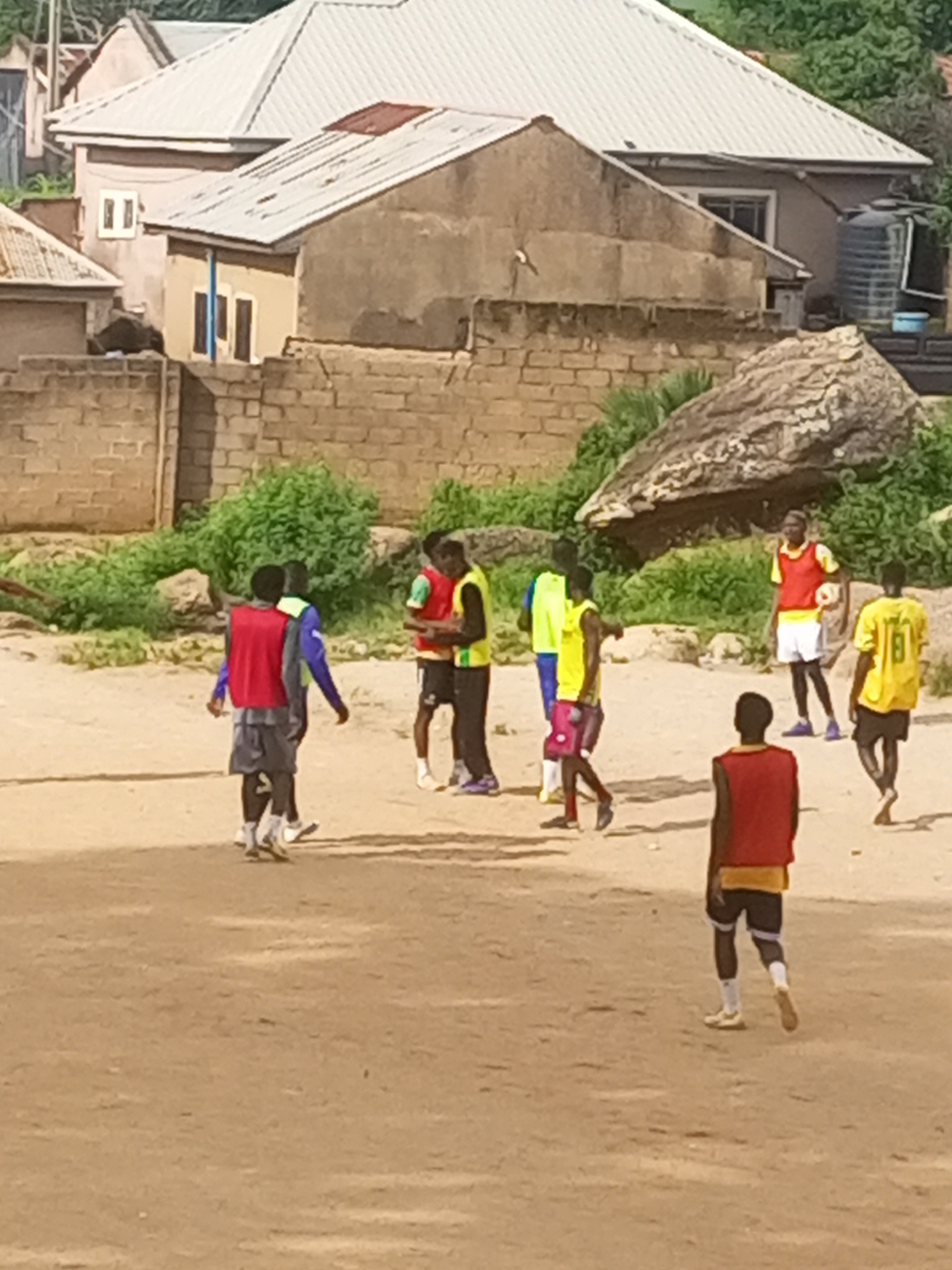 The image depicts a group of young men in athletic attire, appearing to be engaged in a football training session on a dirt field. The scene is set against a backdrop of residential buildings and lush greenery, characteristic of a neighborhood in Jos, Nigeria.
In the foreground, a young man wearing a red vest over a yellow shirt and black shorts is partially visible from the back. To his left, another young man in a red vest and grey shorts is walking away from the camera. Further into the scene, a group of four individuals are clustered together. One of them, in a yellow vest and white shorts, is holding a football. Another in a yellow vest and red shorts is looking towards the viewer. Two other young men, one in a red vest and black shorts, and another in a green shirt and black shorts, are also present in this group.
The environment suggests a somewhat undeveloped area, with the dirt field possibly serving as a local sports ground. The buildings in the background are of a typical Nigerian residential style, with corrugated iron roofs and brick or concrete construction. The presence of large rocks and overgrown vegetation hints at the natural landscape of Jos. The lighting indicates it is daytime, likely with moderate sunlight. The overall atmosphere is one of casual sporting activity within a community setting.