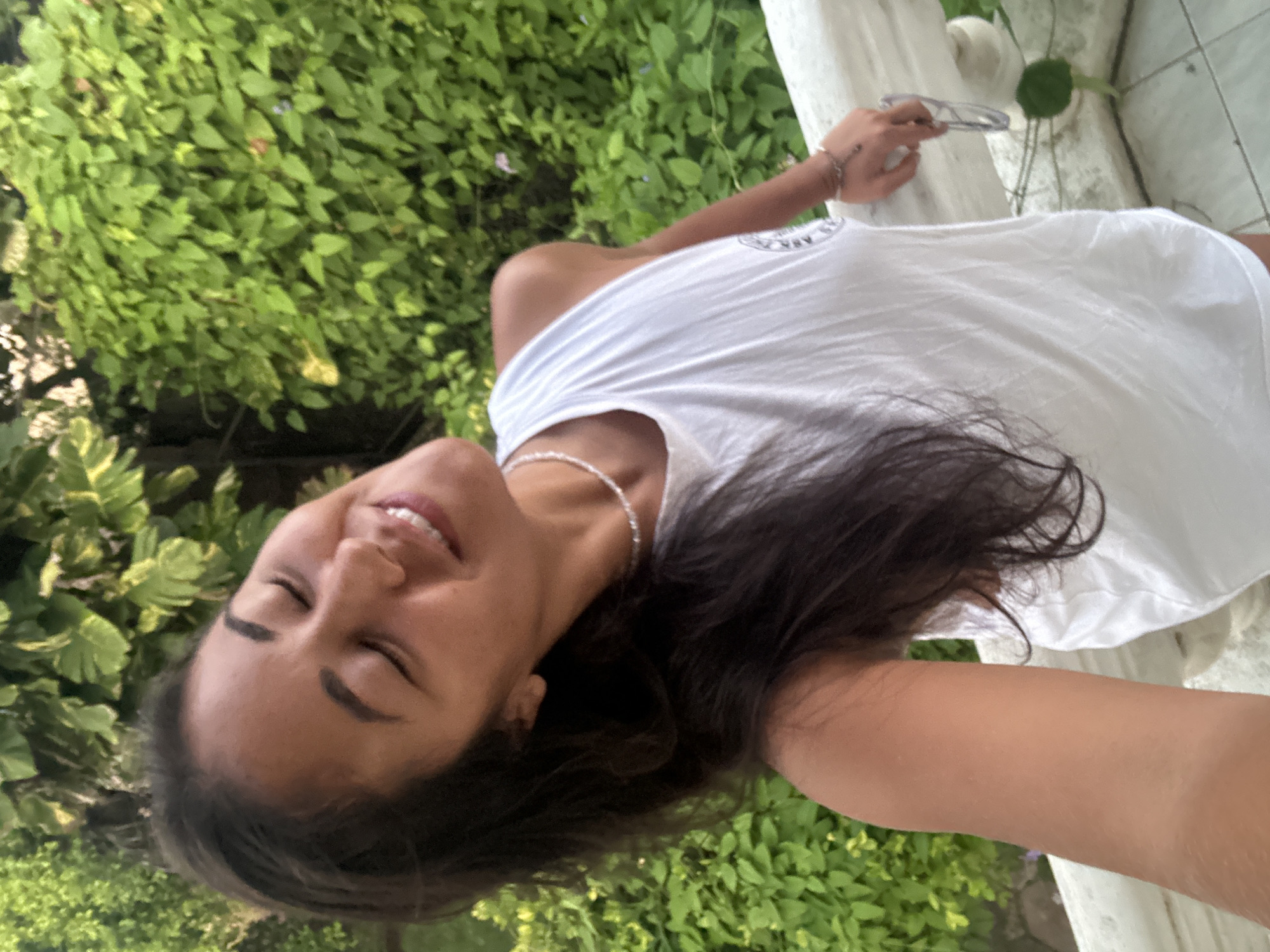 A woman with dark hair is smiling with her eyes closed, tilting her head back, and looking up. She is wearing a white sleeveless top and a beaded necklace. Her right arm rests on a white railing, and she is holding a pair of glasses in her hand. She appears to be outdoors, with lush green foliage in the background. The setting suggests a tropical environment, possibly a balcony or terrace. There is no visible indication of the specific location within Peleana, Sri Lanka.