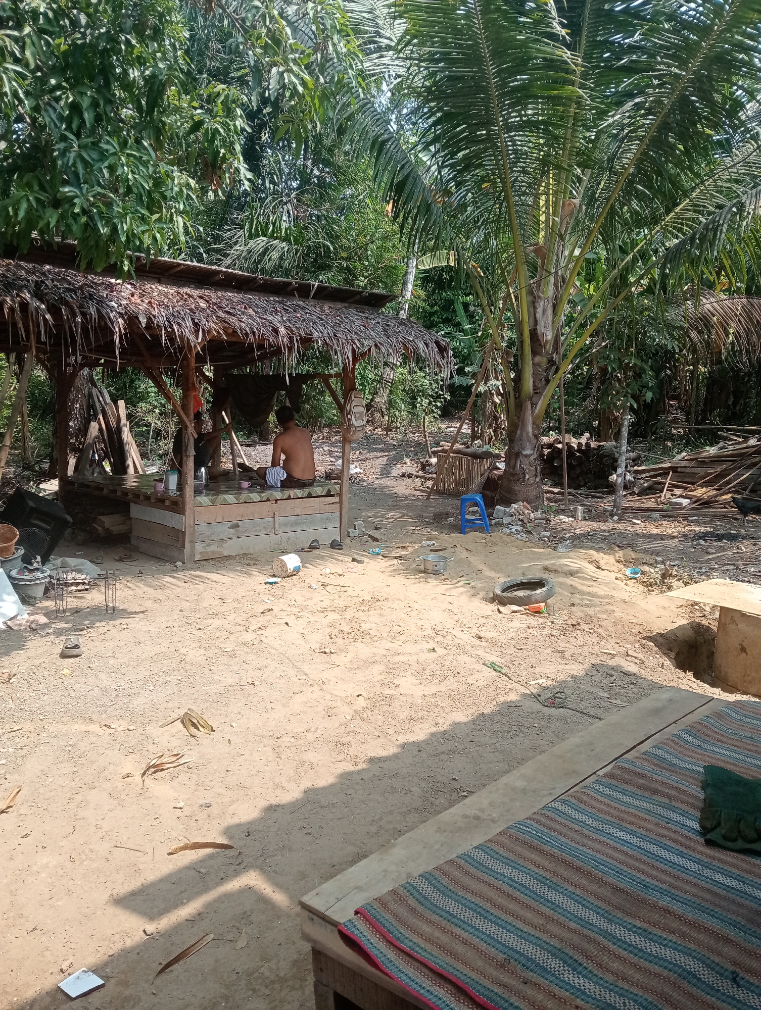 The image depicts an outdoor scene in Muara, Indonesia. In the center of the frame, a man is seated within a rustic shelter constructed with a thatched roof and wooden supports. He appears to be engaged in an activity, possibly preparing food or working on a craft, as indicated by items on the platform he is on. The shelter is elevated and surrounded by a sandy ground. To the left of the shelter, various objects are scattered, including buckets and what appears to be building materials. In the background, dense foliage and palm trees are visible, suggesting a tropical environment. The ground is a mixture of sand and dry leaves, with a tire and a blue plastic stool also present in the vicinity. A woven mat with striped patterns is laid out in the foreground, suggesting an area for resting. The overall impression is of a simple, rural dwelling or workspace in an Indonesian setting.