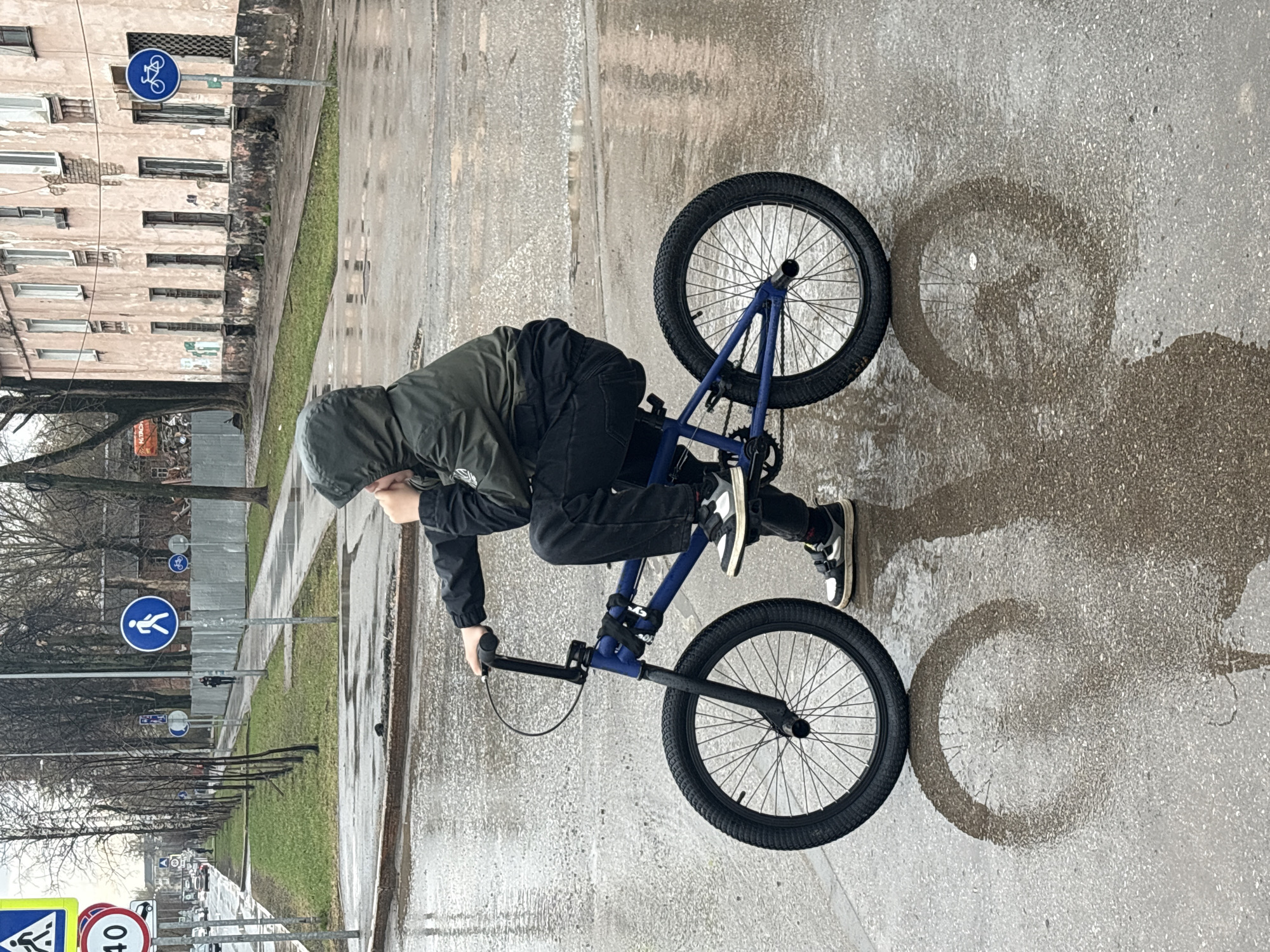 A person wearing a hooded jacket and sneakers is balancing on a blue BMX bike in Perm, Russia. The person is positioned on a wet sidewalk, with the bike's wheels reflecting on the wet surface. The background features a street with buildings, trees, and street signs, including signs for pedestrian and bicycle routes. The scene appears to be overcast or rainy.