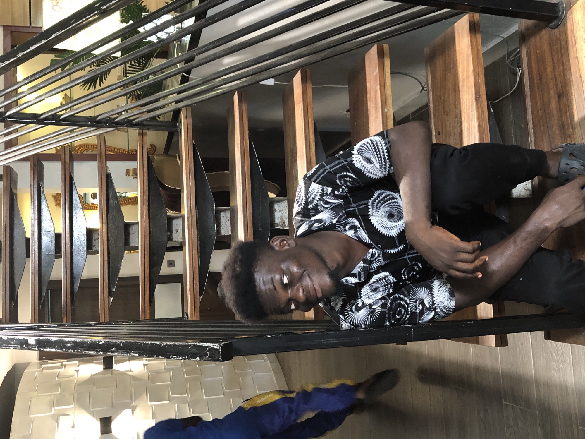 The image, rotated 90 degrees counter-clockwise, displays a young Black man posing on a modern indoor staircase. He is positioned on a wooden step, leaning against a black metal railing composed of multiple horizontal bars. The man wears a black and white patterned short-sleeved shirt with intricate circular designs, black pants, and dark clogs or sandals. He has short, dark hair and a beard, and looks directly towards the viewer with a slight smile, his left arm resting across his body.
The staircase features warm-toned wooden treads and risers, contrasting with the dark metal railing. Through the balusters, elements of the background are visible, including light-colored walls, some golden decorative accents, and green foliage from a large plant. Below the man, the lower body and feet of another individual, dressed in blue and yellow pants, are partially seen on a light-colored, possibly tiled or reflective floor. The scene suggests a well-lit contemporary interior, likely within a building with multiple levels. No visible text is present.