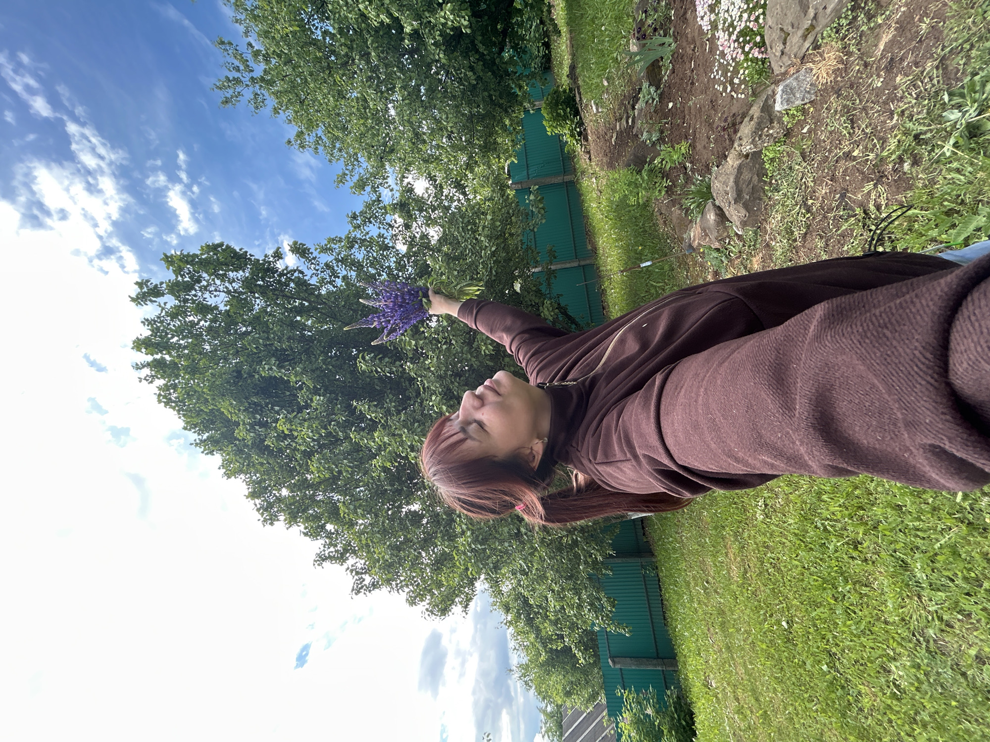 A young woman is lying in a grassy area holding a bouquet of purple lupines. She is wearing a brown sweatshirt. Behind her are trees and a blue sky with scattered clouds. A green fence is visible further back, and a garden bed with rocks and flowers is in the background. It is daytime, and the location is Фоки, Russia.