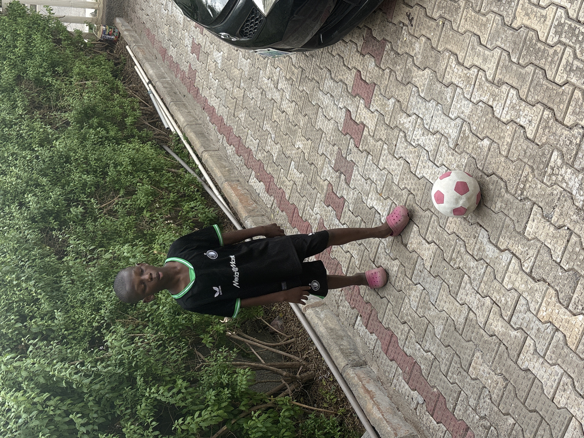 A young male stands on a brick-paved surface adjacent to a lush green hedge. He wears a black soccer jersey with green trim, black shorts, and pink Crocs. A soccer ball with pink pentagonal patterns lies on the pavement near his feet. A portion of a black car is visible in the background. The location is Iddo Tudunwada, Nigeria.