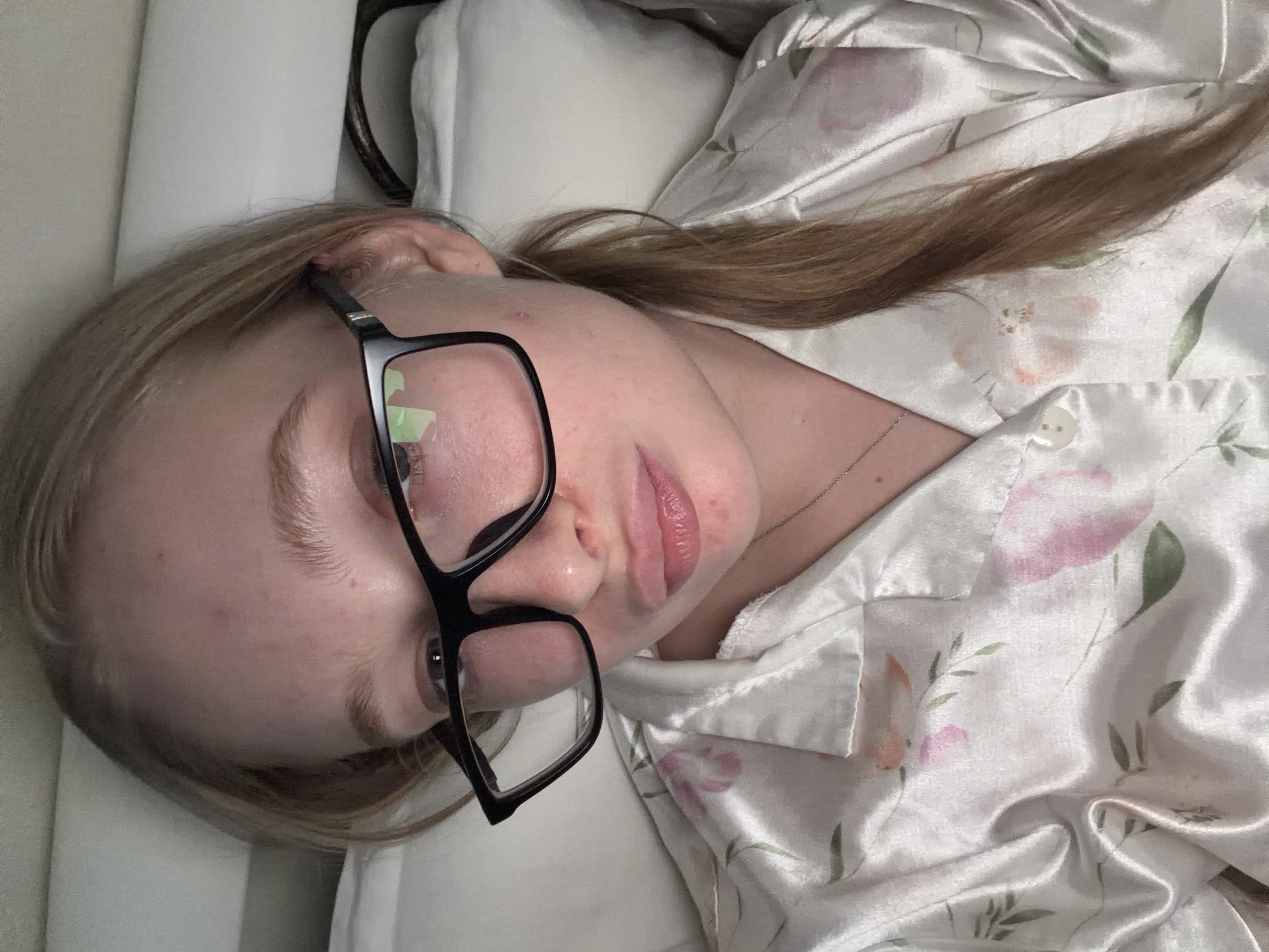 A woman lies in bed, wearing black-framed glasses and a white floral pajama top. She is positioned on a white pillow. The setting appears to be indoors. The location information is not discernible from the image.