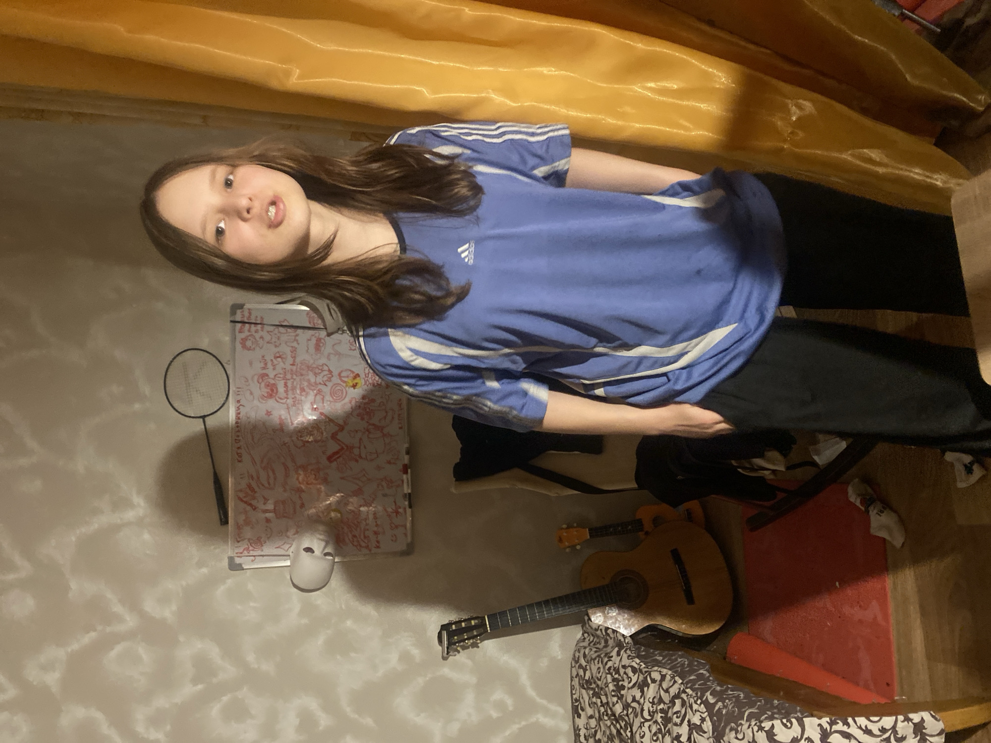 A young female with long brown hair stands in a room. She is wearing a blue Adidas t-shirt with white stripes and black pants. Her hands are in her pockets. Behind her, there is a whiteboard with drawings, a badminton racket, a guitar, and a bed with a patterned comforter. To her right, there is a yellow curtain. The room appears to be a bedroom, likely in an apartment, given the interior setting and personal belongings visible. The lighting suggests it is likely nighttime or an interior space with artificial lighting. The presence of personal items such as musical instruments and artwork on the whiteboard indicates a lived-in, personal space. The overall impression is a candid, informal moment captured within a domestic setting.