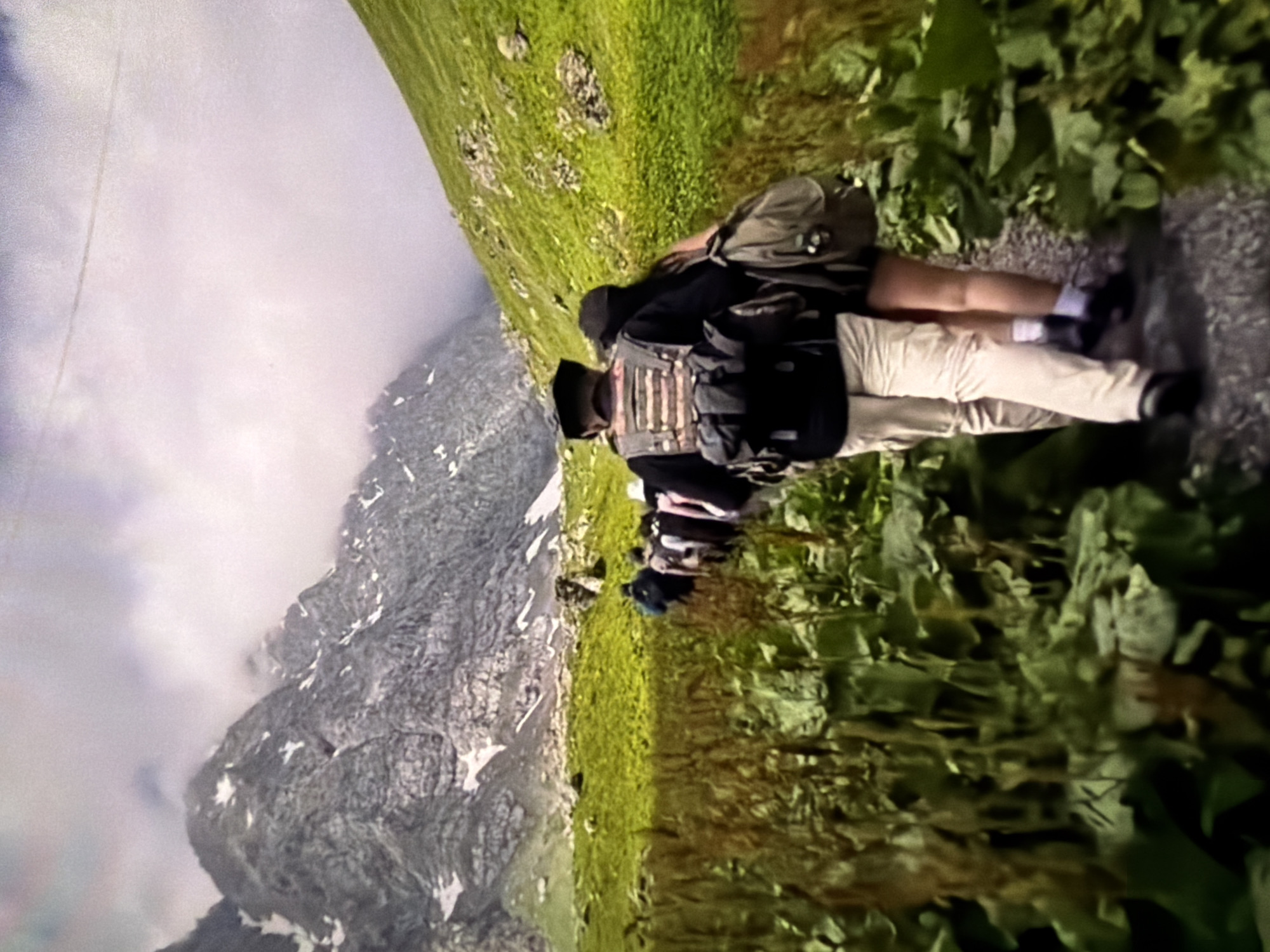 The image captures a scene in a mountainous region of Unknown, Unknown. A person is situated on a path or trail, wearing a dark top, light-colored pants, and black shoes with white socks. They have a backpack and what appears to be a vest or harness. The person is positioned between a patch of dense green foliage to one side and a grassy slope on the other. In the background, a towering mountain is partially obscured by clouds, displaying rocky faces and patches of snow.