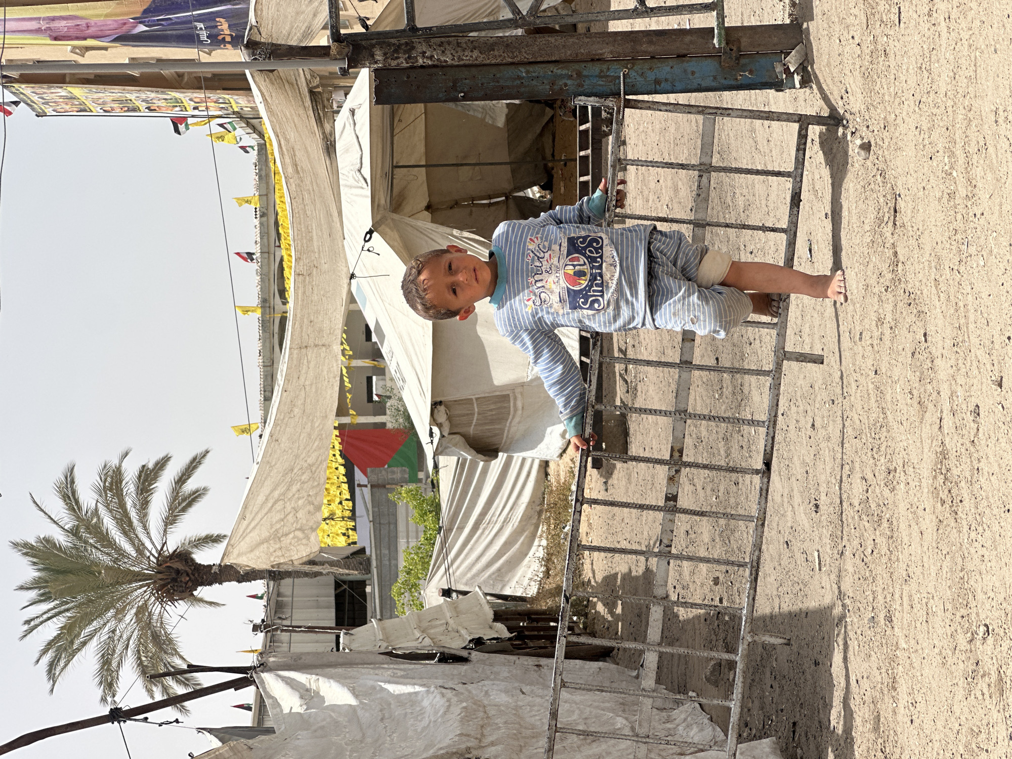A young barefoot child, with short dark hair and wearing a blue and white striped long-sleeved shirt and pants with a "Smiles" graphic on the shirt, stands on a weathered metal gate or fence. The child appears to be looking directly at the camera. The background features a makeshift camp or settlement with white tents and various banners and flags, including Palestinian flags, strung above. A tall palm tree stands to the left of the tents. The ground is dusty and sandy. The scene is likely located in the city of Gaza, Palestinian Territories, based on the flags and camp-like environment.