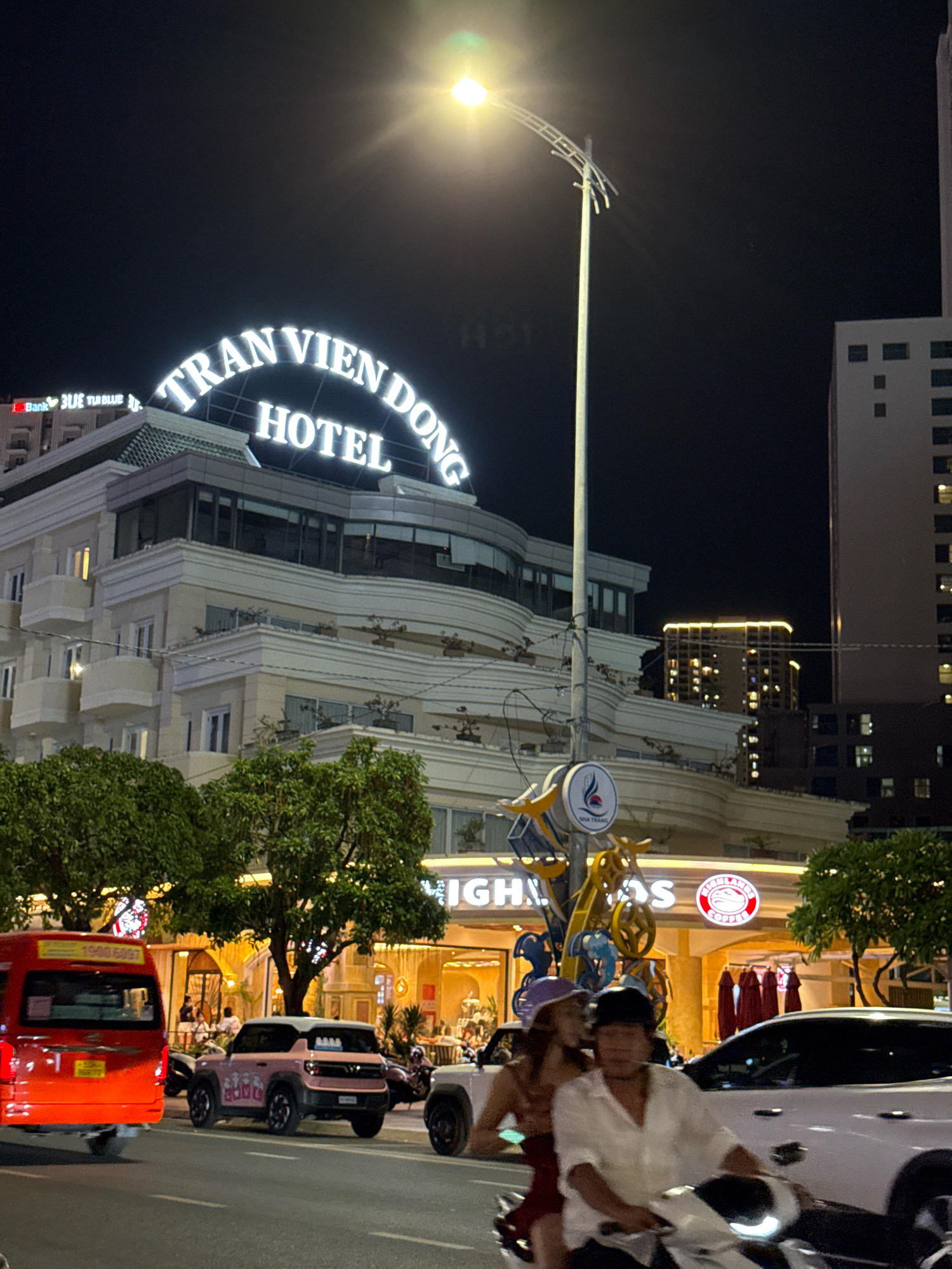 In Nha Trang, Vietnam, at night, a wide street is captured, featuring a prominent hotel with a large illuminated sign that reads "TRAN VIEN DONG HOTEL". Adjacent to the hotel, a bank sign is visible with the text "Bank BLUE TUDblue". On the street, various vehicles are present, including a bright orange bus on the left, a light pink SUV, and several white cars and motorcycles. Two individuals are riding on a motorcycle in the foreground, with a woman wearing a purple hat and a man in a white shirt. The background displays a brightly lit building with illuminated windows and trees. A street lamp with a bright light source is also visible.