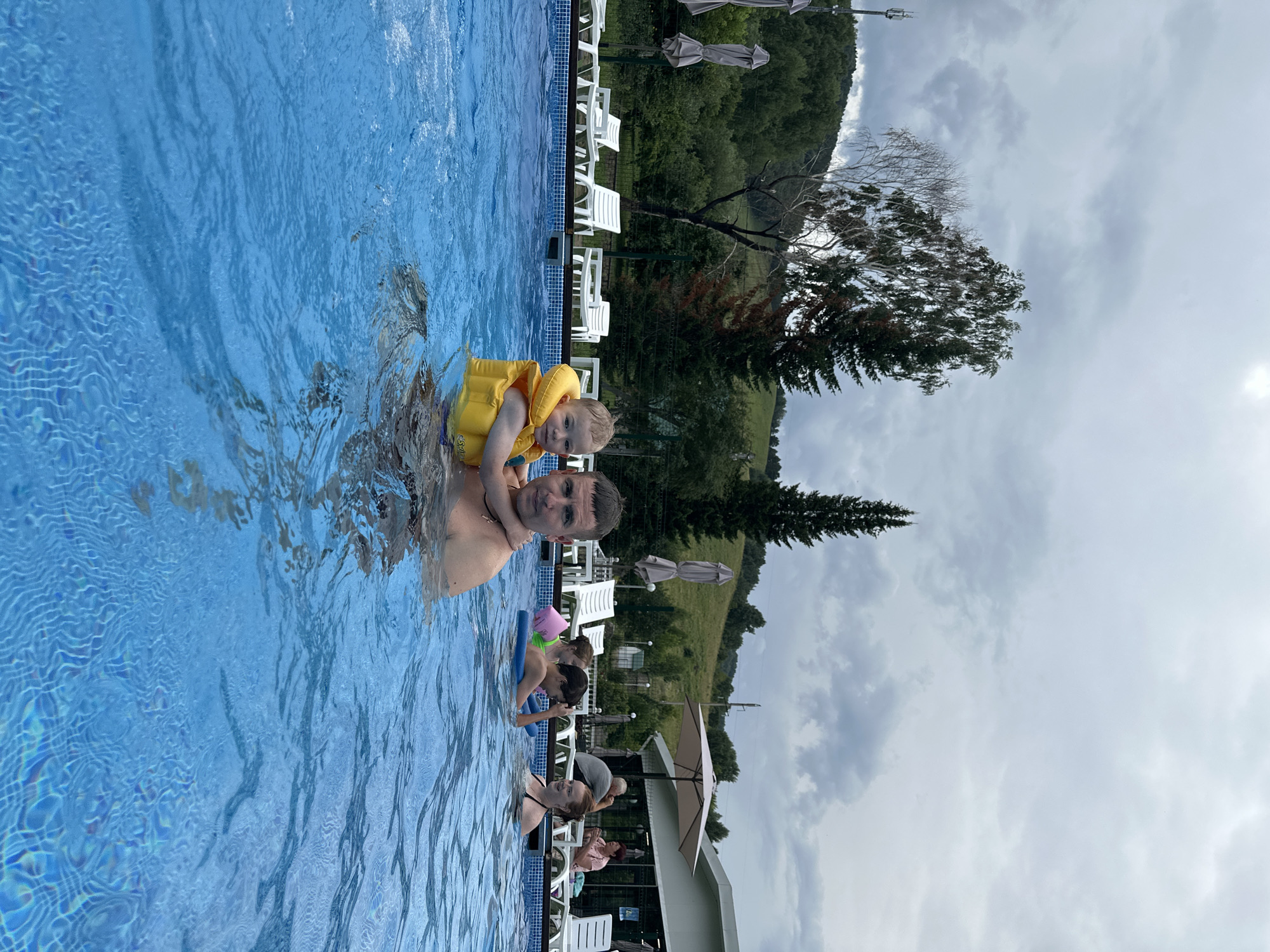 A low-angle medium shot captures a father and his young child in a blue swimming pool, with the child wearing a yellow life jacket. They are positioned in the foreground, with the father's head and shoulders visible, and the child being held by him. Other people are in the pool in the background, along with lounge chairs, umbrellas, and a building. The environment suggests an outdoor swimming pool area. The sky is overcast with heavy clouds. The location is the city of Даниловка (алтайский), Russia.