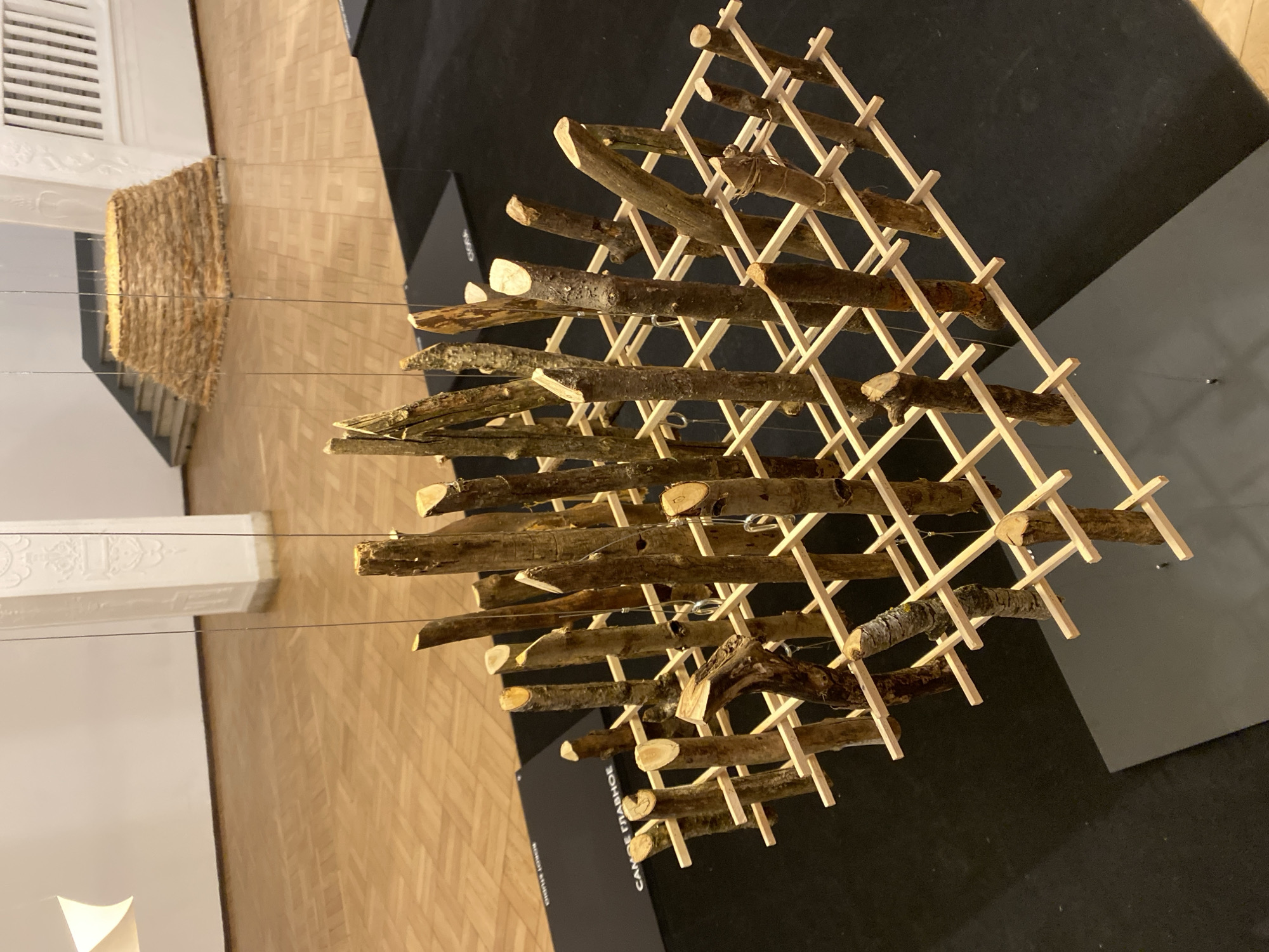 The image captures an art installation inside a building, likely a museum or gallery. The installation appears to be a suspended three-dimensional structure constructed from wooden sticks. A framework of light-colored thin wooden pieces forms a grid-like pattern, holding a collection of natural, darker-colored wooden branches or twigs. The structure is suspended from the ceiling by thin wires. The background includes parquet flooring, a section of a white wall with architectural details, and what seems to be another art piece made of straw or similar material. A black platform or display stand is also visible, with writing on the side, although the details of the text are illegible. The scene is well-lit, creating a clean and modern aesthetic. The location is in the city of Unknown, Unknown.