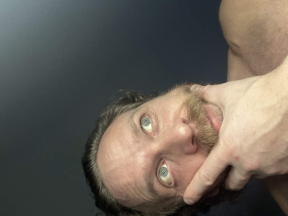 A man with light skin, a light beard, and striking blue-green eyes looks into the camera. His hand, with visible veins, is near his mouth, potentially touching his lips. The background is a muted gradient from light to dark gray. The man's expression is difficult to discern. The photo is tilted, giving an off-kilter perspective. Part of his bare shoulder is visible in the upper right corner.